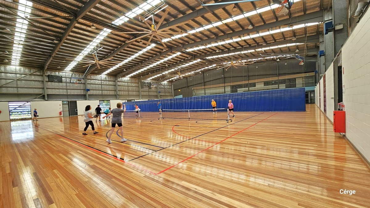Another view of the court. The area is well lit by the long strips fluorescent bulb from the ceiling. The ceiling is painted brown and has corrugated finish, accented by a couple of large brown fans. The timber floor has markings intended for different types of ball games. The far end shows a blue cloth divider. There are numerous people playing badminton. A white table and white bench chairs are also positioned on the left.