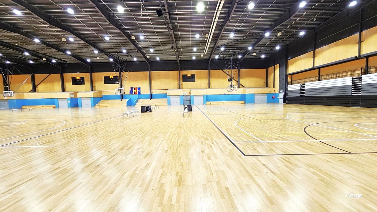 Court 1 is a large and open space and is well-illuminated by fluorescent lights in a mesh-designed ceiling. The timber flooring resembles light brown wood. The space is divided into three sections by tables and chairs. The far wall is a combination of yellow and blue, adorned with multiple flat-screen televisions. Each section has a multi-tiered grandstand for tired seating, while the left wall displays white and brown vertical designs. Additionally, a larger flat-screen monitor is in view.