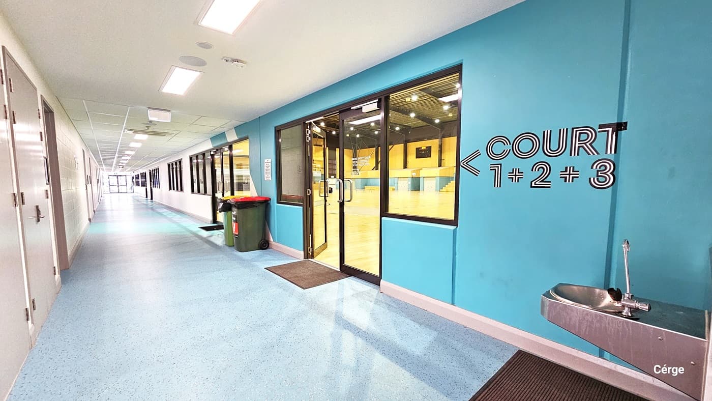 The lengthy lobby features a granite-finished floor and a white ceiling. The right wall is powder blue with signage reading "Court 1, 2, 3" and an arrow pointing forward. Close to the wall is a drinking fountain and a non-slip brown mat underneath. On the left, an open door and window lead to a brightly lit yellow court with two rubbish bins. The left wall is white and has doors to other rooms.