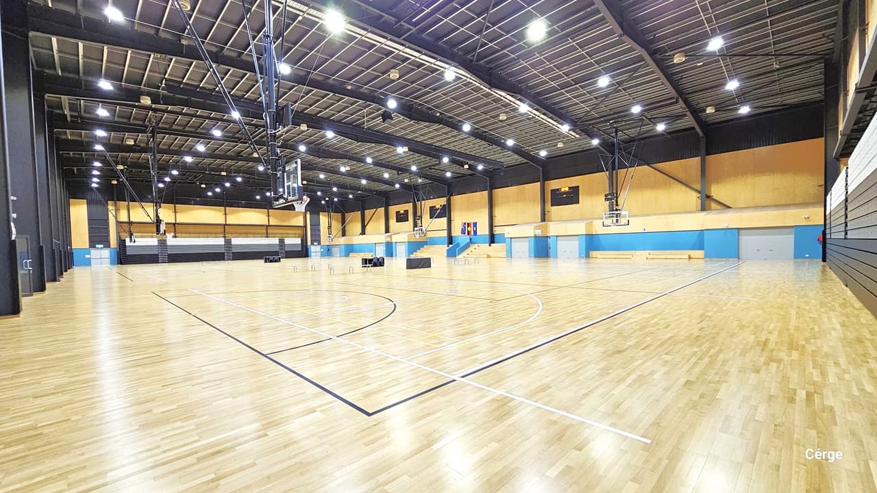 Court 1 is a large and open space and is well-illuminated by fluorescent lights in a mesh-designed ceiling. The timber flooring resembles light brown wood. The space is divided into three sections by tables and chairs. The far wall is a combination of yellow and blue, adorned with multiple flat-screen televisions. Each section has a multi-tiered grandstand for tired seating, while the left wall displays white and brown vertical designs. Additionally, a larger flat-screen monitor is in view.