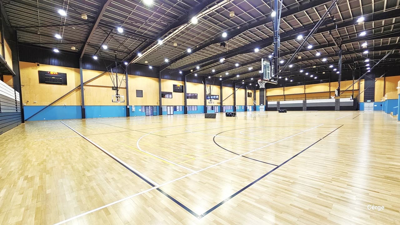 Court 1 is a large and open space and is well-illuminated by fluorescent lights in a mesh-designed ceiling. The timber flooring resembles light brown wood. The space is divided into three sections by tables and chairs. The far wall is a combination of yellow and blue, adorned with multiple flat-screen televisions. Each section has a multi-tiered grandstand for tired seating, while the left wall displays white and brown vertical designs. Additionally, a larger flat-screen monitor is in view.