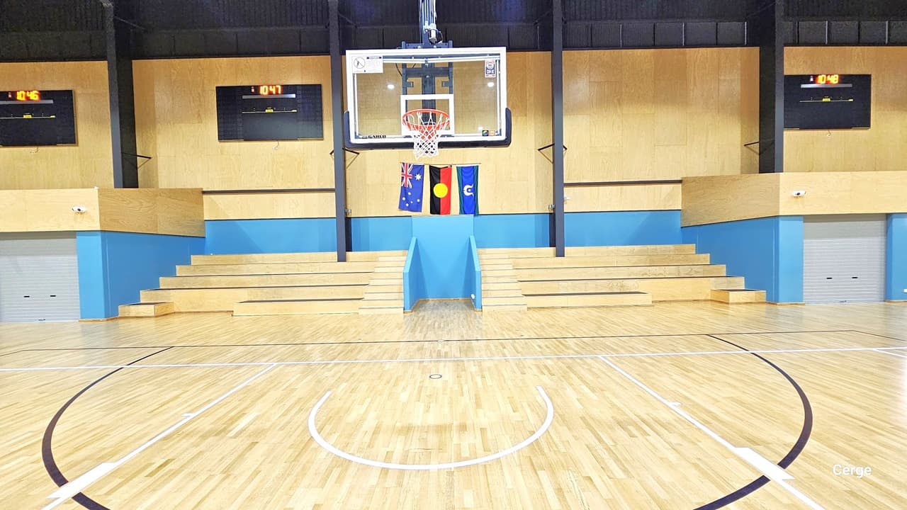 The basketball area features a wooden design timber floor marked with a shooting area in a half-circle. The wall is painted yellow with blue accents, and three flat screens display the time. There's a glass backboard with an orange hoop and white net. Three flags are hung on the wall. This area also includes multi-tier seating.