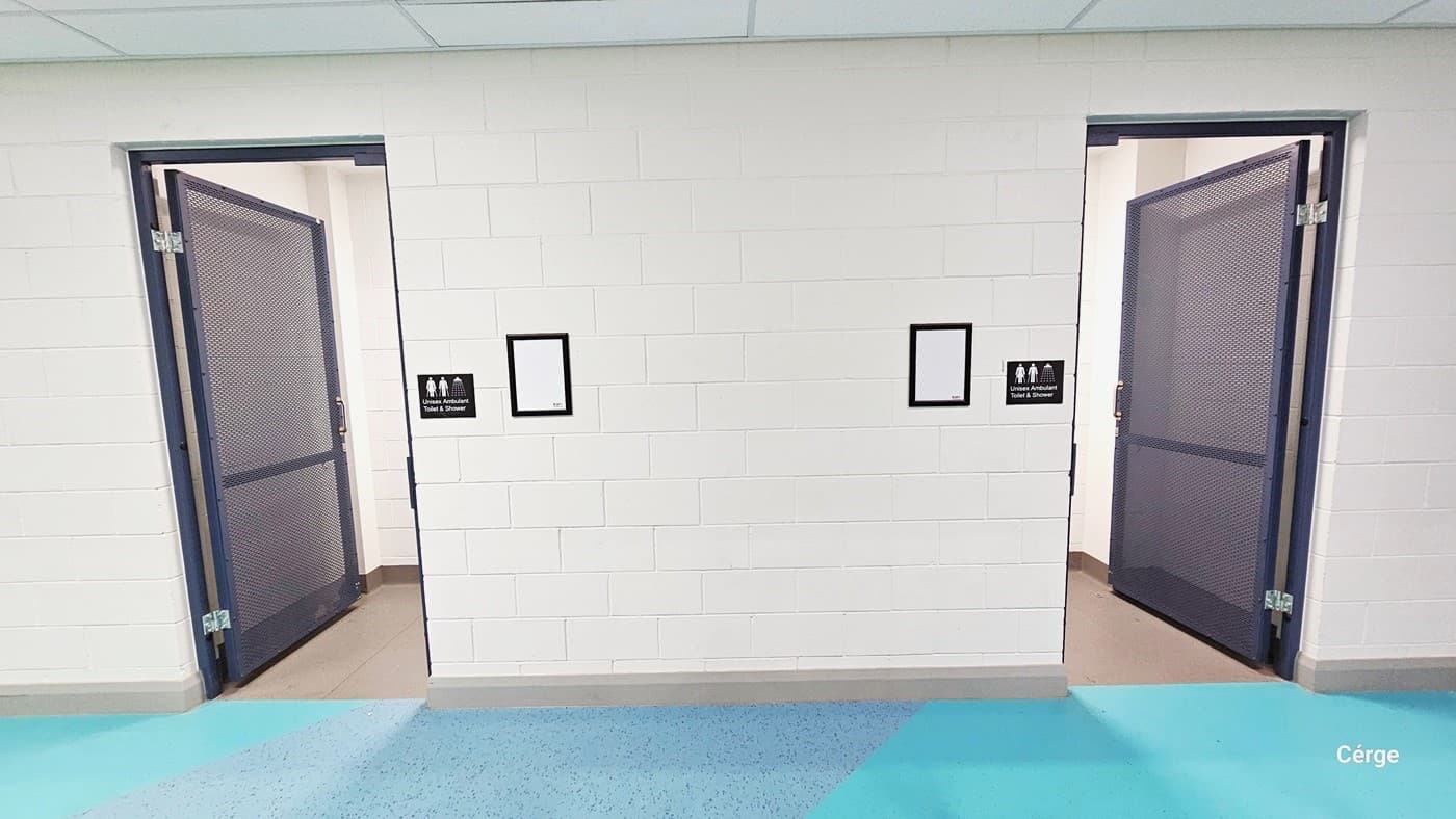 The accessible bathroom is characterized by its white walls with a brick-like design. It features two stainless steel doors with a brown mesh-like pattern, each equipped with a metal handle. Signs on both sides of the doors clearly indicate that this bathroom is gender-neutral, and includes a shower room. Positioned beside the signage are two frames with brown borders. The floor has decor with light green and grey granite-style designs and white ceilings.