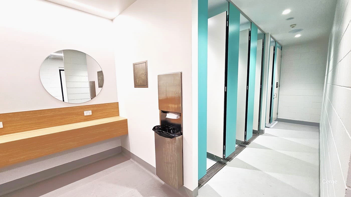 Inside the bathroom, there's a round mirror and a long brown wooden panel on the left side. Adjacent to them are a tissue dispenser, a silver box, and a rubbish bin. The floor has a matte finish in grey and white tones. On the right, there are five bathroom stalls with green walls and white doors. The ceiling and wall exhibit a white colour with a brick-like design.