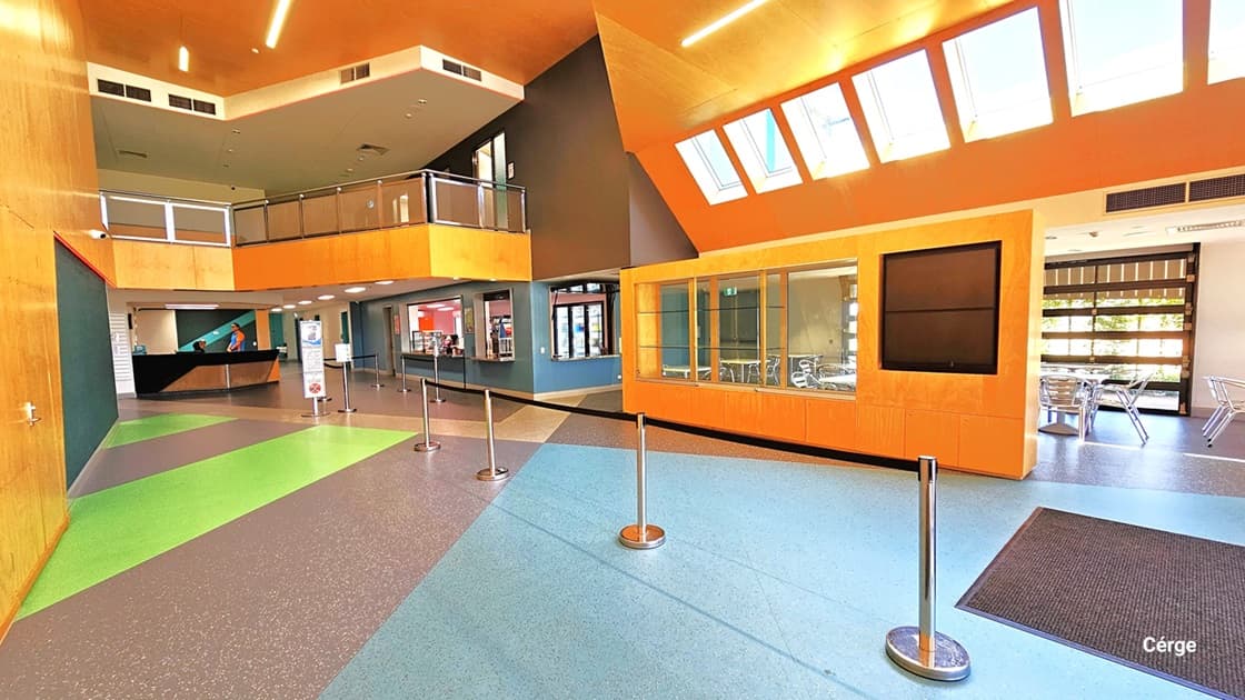 Beyond the entrance, there is a modern orange and white ceiling. The multi-colored floor features stainless steel bollards and signage. To the right, a brown anti-slip mat rests before an open door leading to a cafe. The cafe's orange-painted wall boasts large glass windows, tables, and chairs. Further ahead is another cafe. On the left, a yellow and green wall guides the reception area, offering a glimpse of the second-floor section enclosed by glass railings.