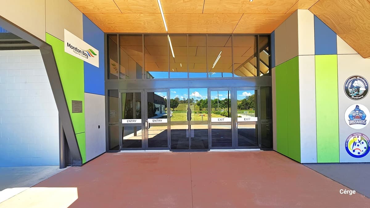 At South Pine Entrance, a glass door with stainless steel handrails serves as the gateway. Two doors, "Entry" on the right and "Exit" on the left, lead the way. The doors reflect the grassy surroundings. The building's floor features faded brown tiles, while the ceiling displays a yellow tile design. A "Moreton Bay" sign is positioned on the upper left near the ceiling. On the right, three circular advertising signs stand out against the wall, painted with blue, apple green, and white stripes.