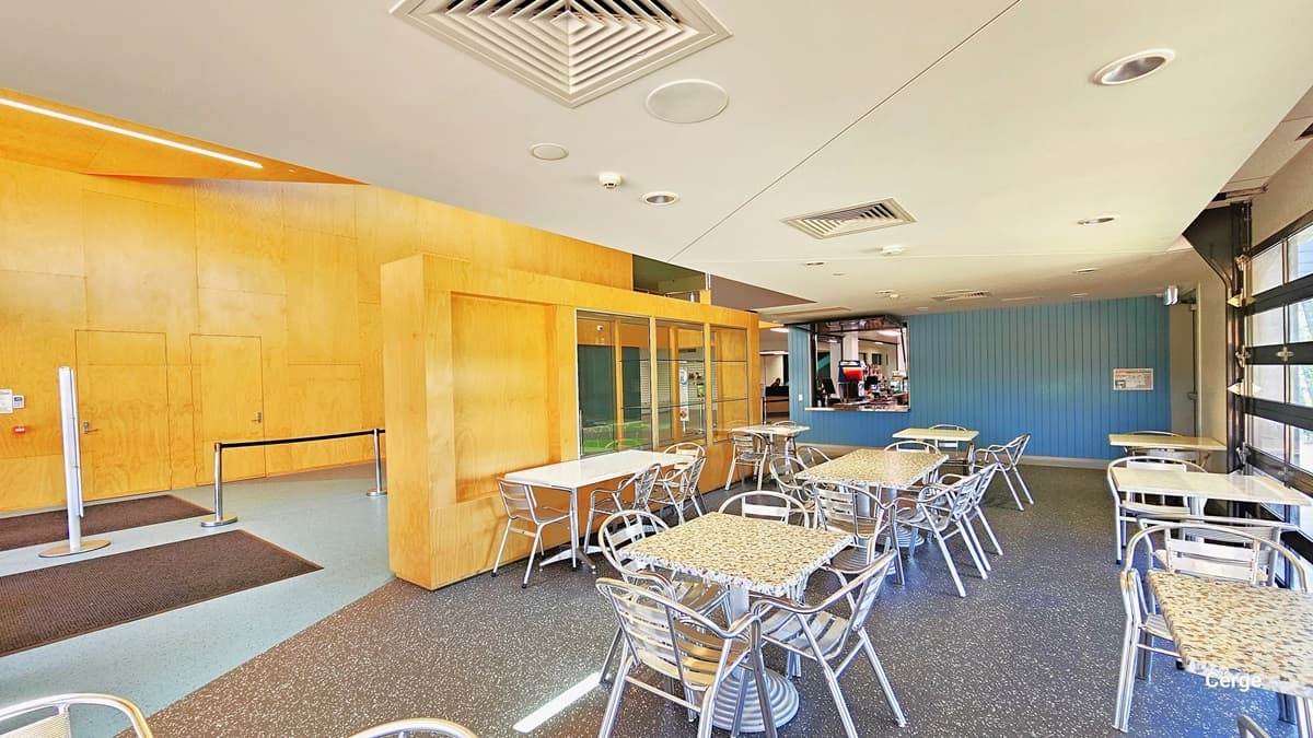 In the cafe, there are well-arranged printed tables and silver stainless steel chairs. The floor displays a granite finish, extending to the area where it meets the blue wall and the cafe's kitchen. The ceiling incorporates two centralized air conditioning systems, fluorescent lights, and a CCTV. The left wall is painted yellow, and the right wall has a large window with brown panels, offering a glimpse of the outside.