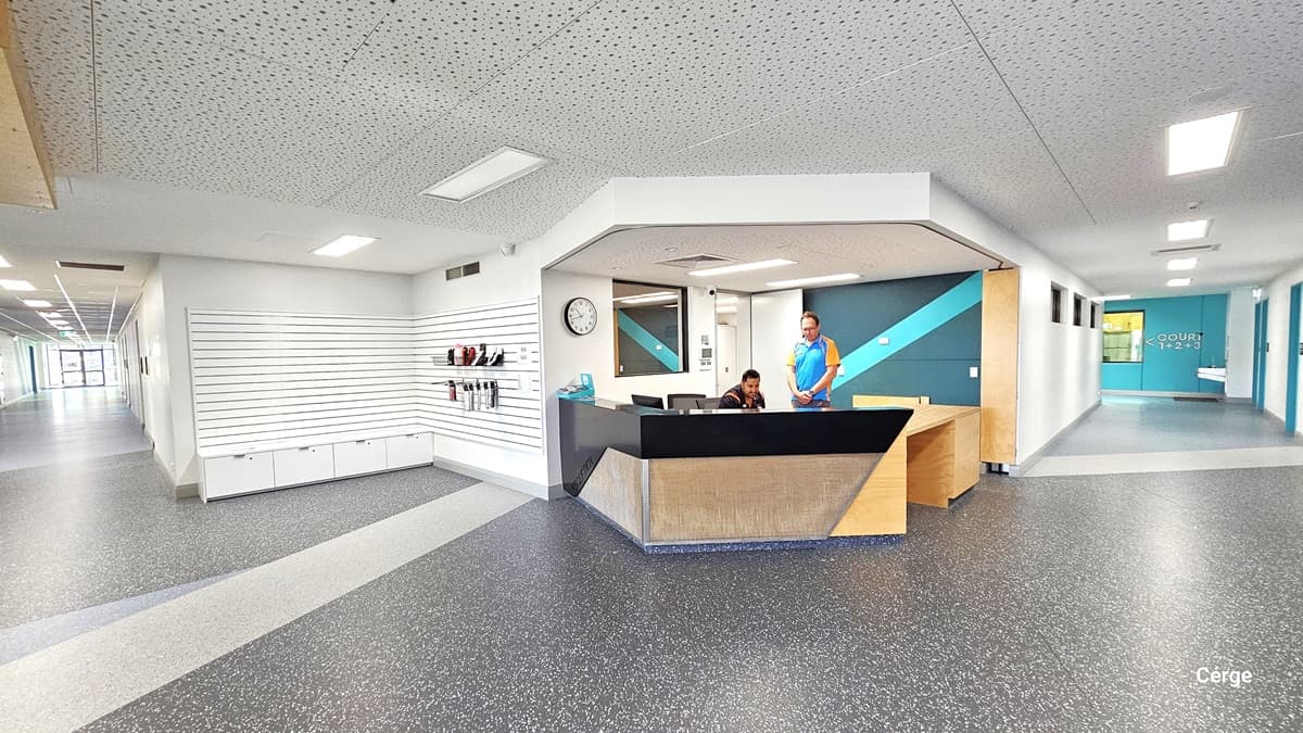 The reception area features a grey granite floor with white stripes and ample lighting. The reception desk has a stylish design with a round clock on the wall. Two staff members are present. On the right, a pathway leads to a green wall, while on the left, another path goes to a well-lit area. To the right, there's a white ribbed wall with merchandise.