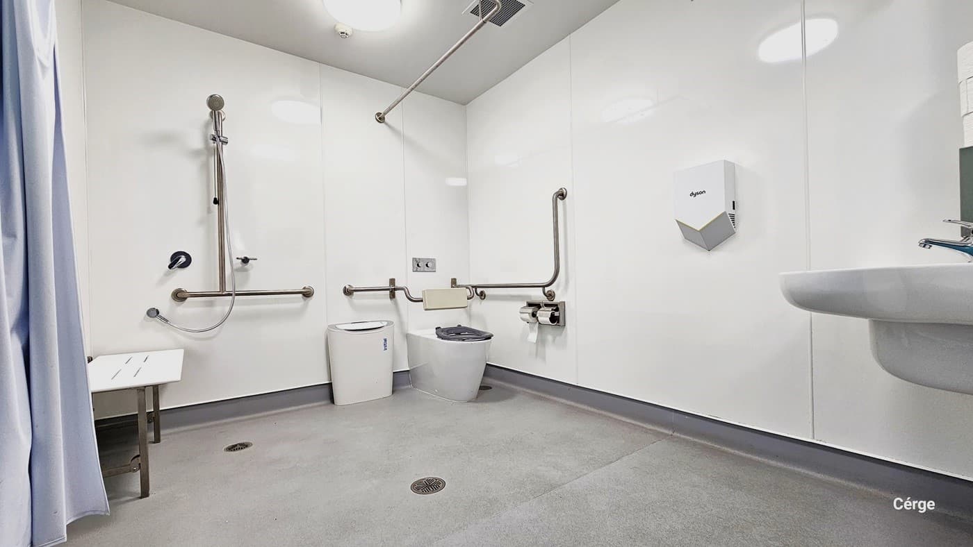 The accessible bathroom within South Pine Indoor Sports is well-lit and spacious, with essential amenities including a functional washbasin, toilet paper holder, rubbish bin, and a bathroom bench. Multiple grab bars and handrails aid mobility. A shower area with a privacy curtain is also available. The grey matte floor complements the bright white walls and ceiling.