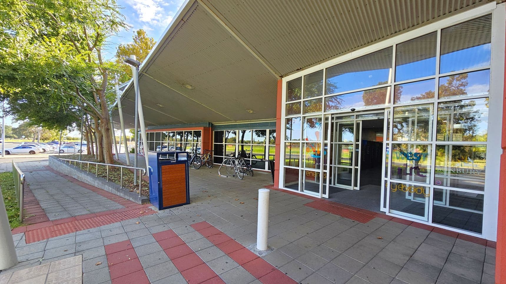 A building entrance with large glass doors and windows on the right. The foreground has a tiled pavement with red and grey patterns. A white bollard and a blue and wooden bin are near the entrance. On the left, there are trees and a row of parked cars. Bicycles are parked under a covered area with a slanted roof. The background shows more trees and a grassy area.