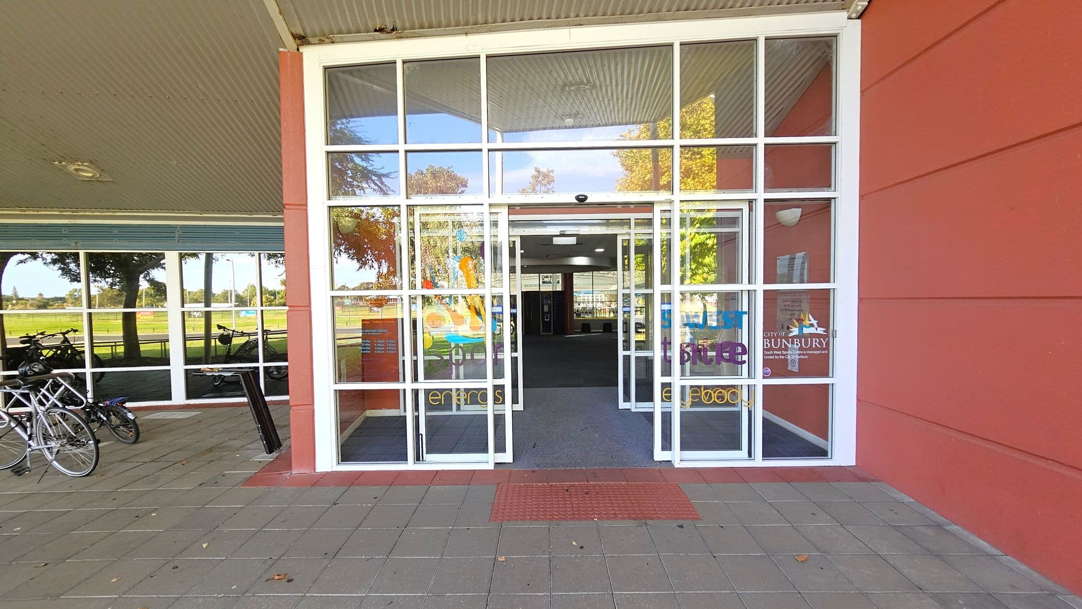 A glass entrance with double doors is centred, featuring colourful text and logos. The building has a red exterior wall on the right. On the left, bicycles are parked against a window reflecting trees and a grassy area. The floor is paved with grey and red tiles, and a red tactile mat is in front of the entrance. The ceiling is corrugated metal with a light fixture.