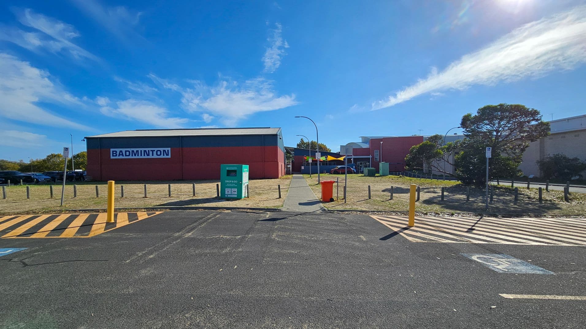 A paved pathway leads to a large red building with a sign reading "BADMINTON" on the left. In the foreground, there is a parking area with yellow and black striped markings and a yellow bollard. A green "DROP & GO" bin and an orange bin are near the path. On the right, a tree and more buildings are visible. The area is bordered by short wooden posts, and a few cars are parked on the left side.