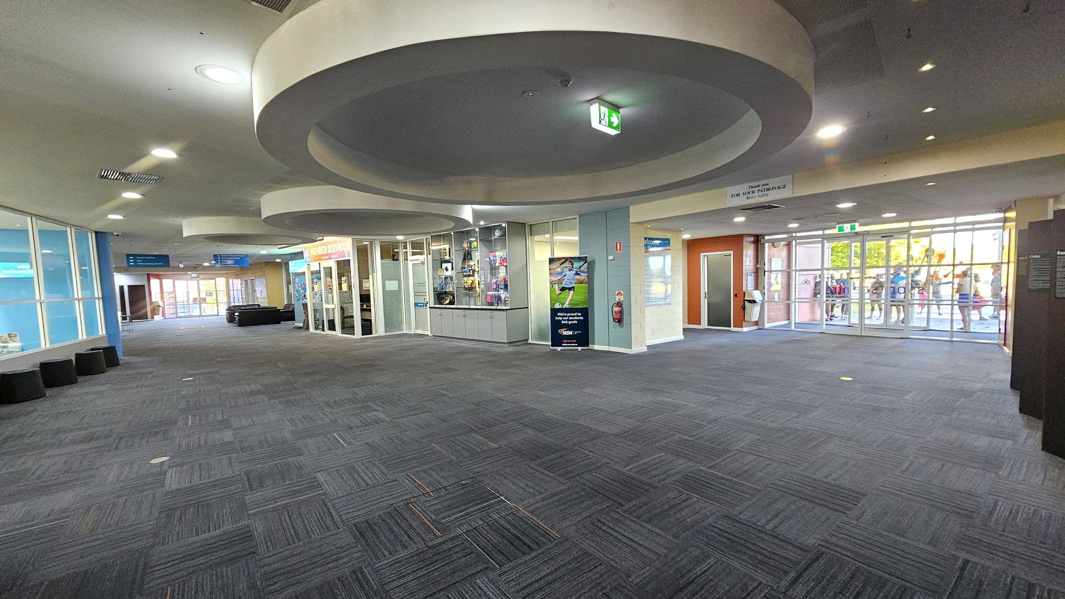 A spacious indoor area with a grey carpeted floor. On the left, there are large glass windows and a row of black cylindrical seats. In the centre, a reception area with glass walls displays various items. Circular ceiling features with recessed lighting are visible above. On the right, glass doors lead outside, where a group of people can be seen. The walls are a mix of light blue and orange tones, and there are signs and posters throughout the space.
