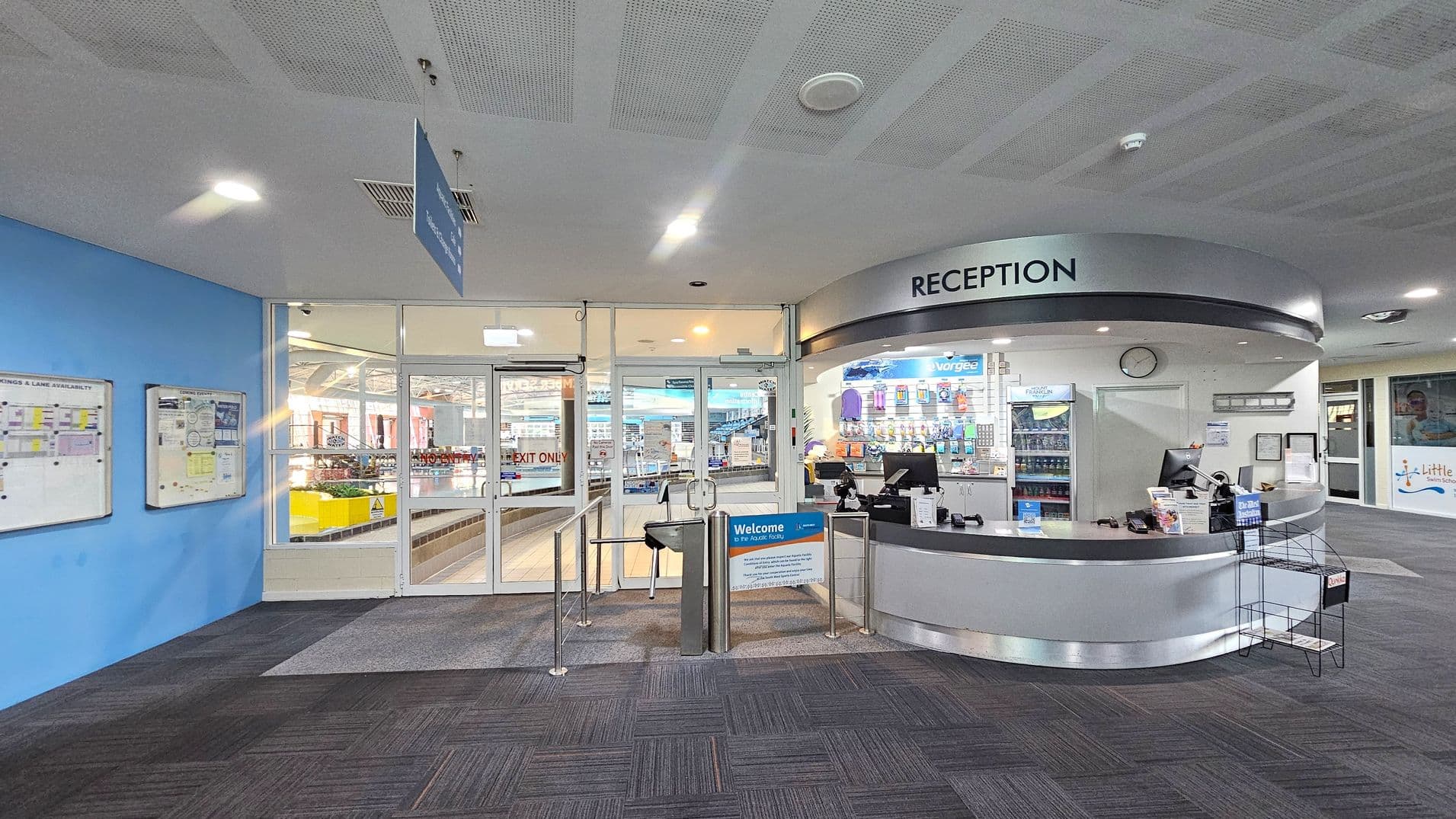 Reception area with a curved desk on the right, featuring computers, brochures, and a display of swim-related products. A sign above reads "RECEPTION". In the centre, glass doors lead to a pool area, with a turnstile and a welcome sign in front. On the left, a blue wall displays notice boards with information. The floor is carpeted in dark grey, and the ceiling is white with recessed lighting.