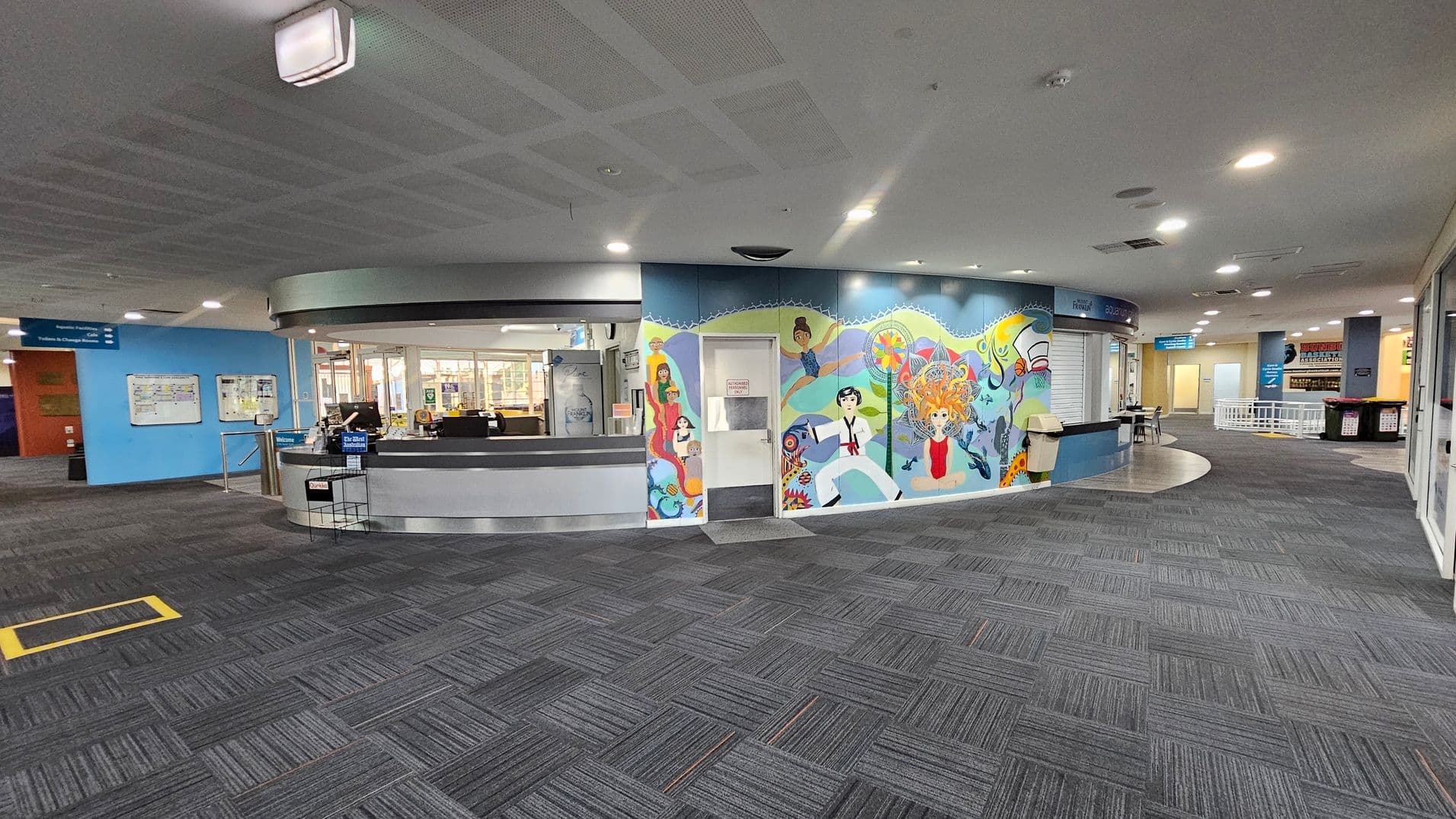 A spacious indoor area with a grey carpeted floor and a ceiling with recessed lighting. On the left, there's a circular reception desk with computers and a "Welcome" sign. Behind it, a blue wall features a colourful mural with various figures and abstract designs. To the right, a corridor leads to other rooms, with signs and a small seating area visible. The overall atmosphere is modern and organised.