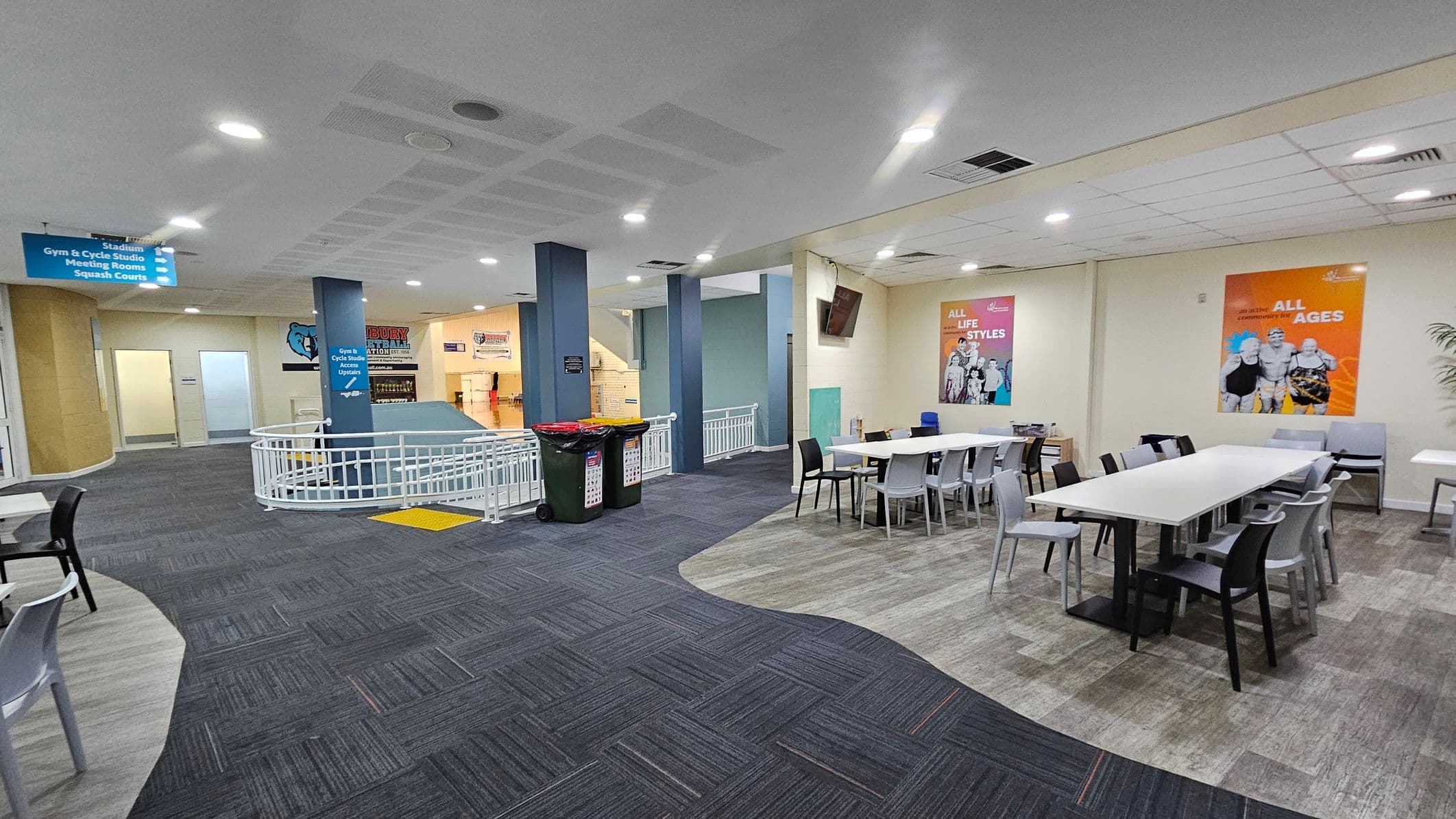 A spacious indoor area with a mix of grey and purple carpeted flooring. On the right, there are white tables surrounded by black and grey chairs, with colourful posters on the wall above. In the centre, a blue pillar with directional signs and a railing around a ramp. On the left, a corridor leads to other rooms, and there are bins near the pillar. The ceiling is fitted with recessed lighting.