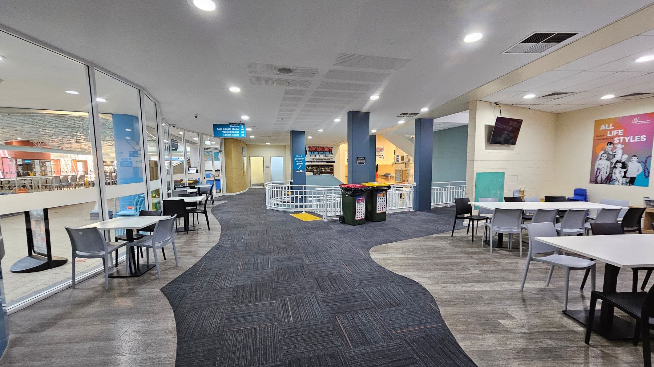 A spacious indoor area with a curved pathway featuring a mix of grey and purple carpet. On the left, there are glass walls with tables and chairs set against them. The right side has more tables and chairs, with a colourful poster on the wall and a mounted TV. In the centre, two bins are placed near a white railing. The ceiling is white with recessed lighting, and there are signs hanging from it. The floor transitions from carpet to a wood-like texture.