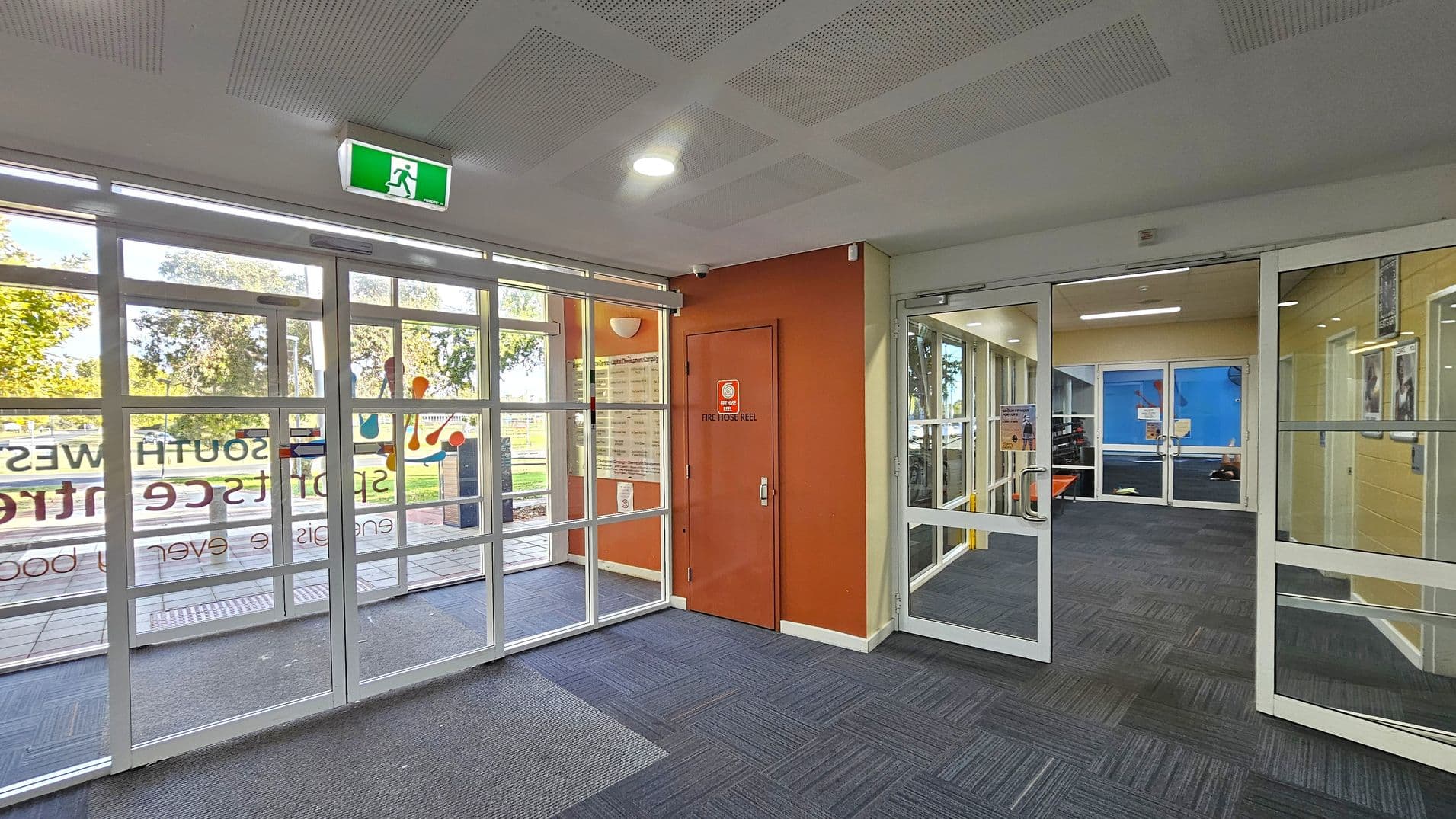 A modern interior space with glass doors on the left, featuring signage and a view of trees outside. In the centre, an orange wall with a door marked "Fire Hose Reel". On the right, more glass doors lead to another room with blue walls. The floor is covered with dark grey and blue patterned carpet tiles, and the ceiling has a grid pattern with recessed lighting. An illuminated green exit sign is above the left doors.
