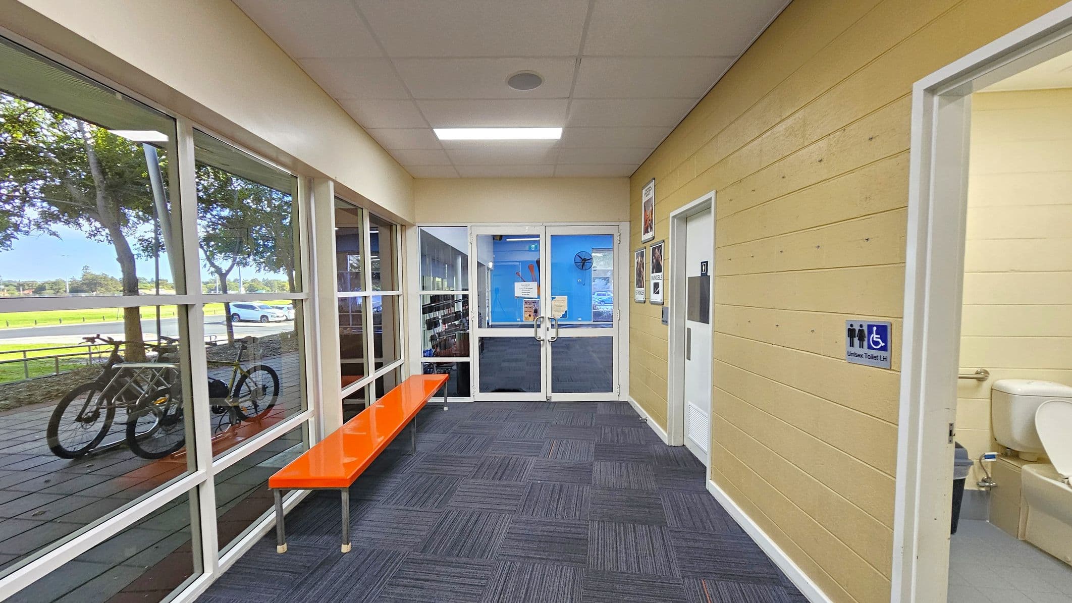 A hallway with a glass wall on the left, showing a view of trees, a parked bicycle, and a car outside. An orange bench is positioned along the glass wall. The right side has a light yellow wooden wall with a door leading to a unisex toilet, partially visible. The floor is covered with blue patterned carpet tiles. At the end of the hallway, there are double glass doors with notices and a blue wall visible through them. The ceiling is fitted with a fluorescent light.