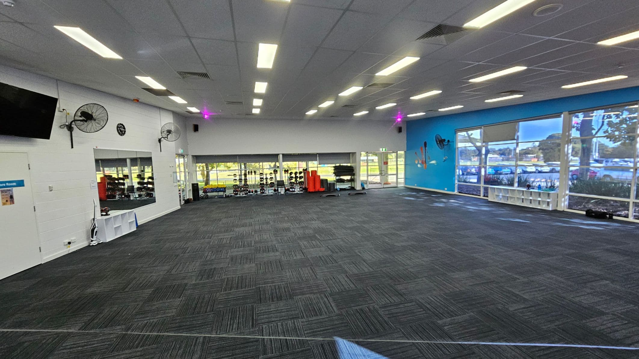 A spacious gym room with a dark carpeted floor and a white ceiling with fluorescent lights. On the left, there's a wall with a mounted TV, two fans, and a clock. Below, a door and a window reveal gym equipment inside. The back wall has large windows showing outdoor greenery and more gym equipment like weights and mats. The right wall is blue with large windows, allowing natural light in. A small shelf is visible beneath the windows.
