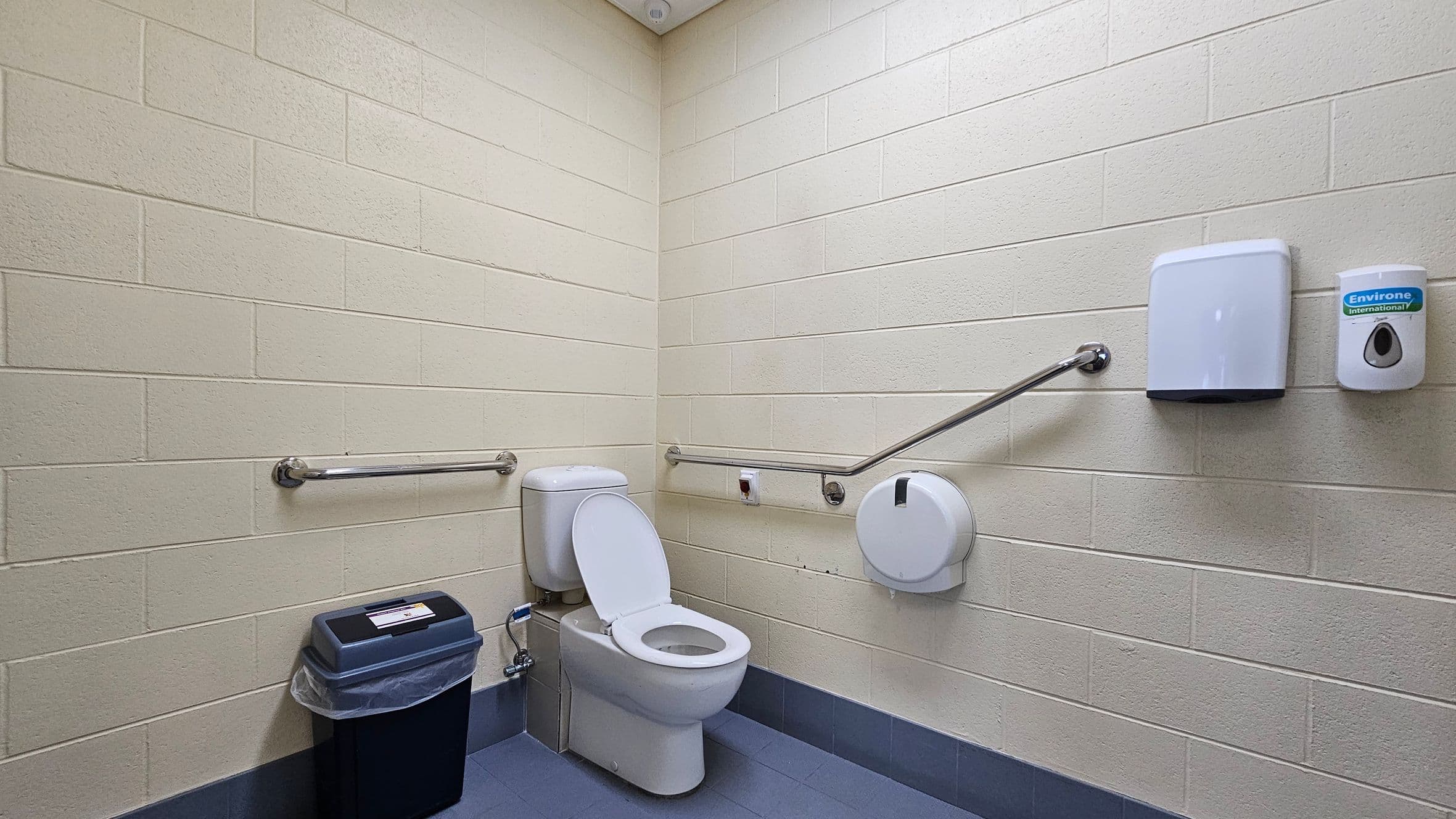 A public restroom with light-coloured brick walls and a blue-tiled floor. In the centre, a white toilet with the seat up is positioned against the corner. To the left, a black bin with a liner is placed on the floor. On the right wall, a toilet paper dispenser, a paper towel dispenser, and a soap dispenser are mounted. Metal grab bars are installed on both walls for accessibility. The ceiling is plain and light-coloured.