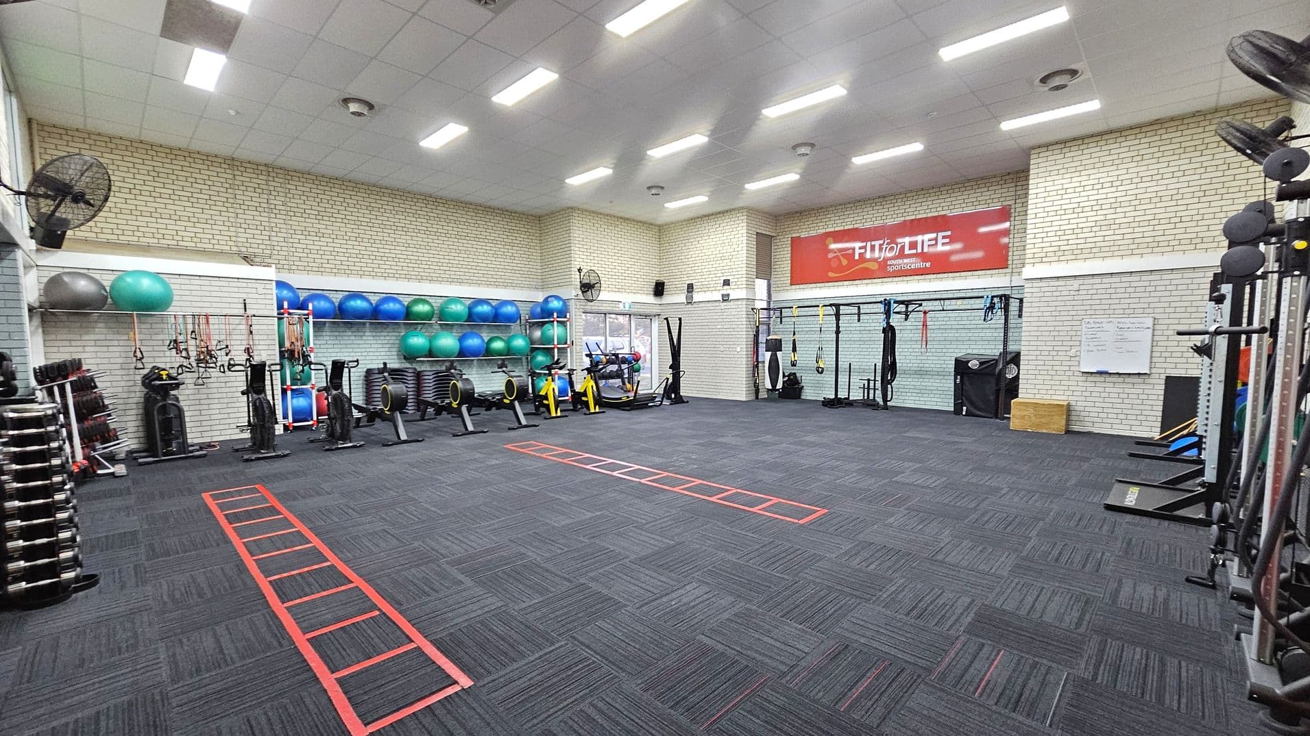 A spacious gym with a dark carpeted floor featuring red agility ladders. On the left, there are stacks of dumbbells, exercise bikes, and a variety of exercise balls on shelves. The right side has more equipment, including resistance bands and a whiteboard. The back wall displays a large red "Fit for Life" banner. The ceiling is fitted with bright fluorescent lights, and the walls are made of light-coloured bricks.