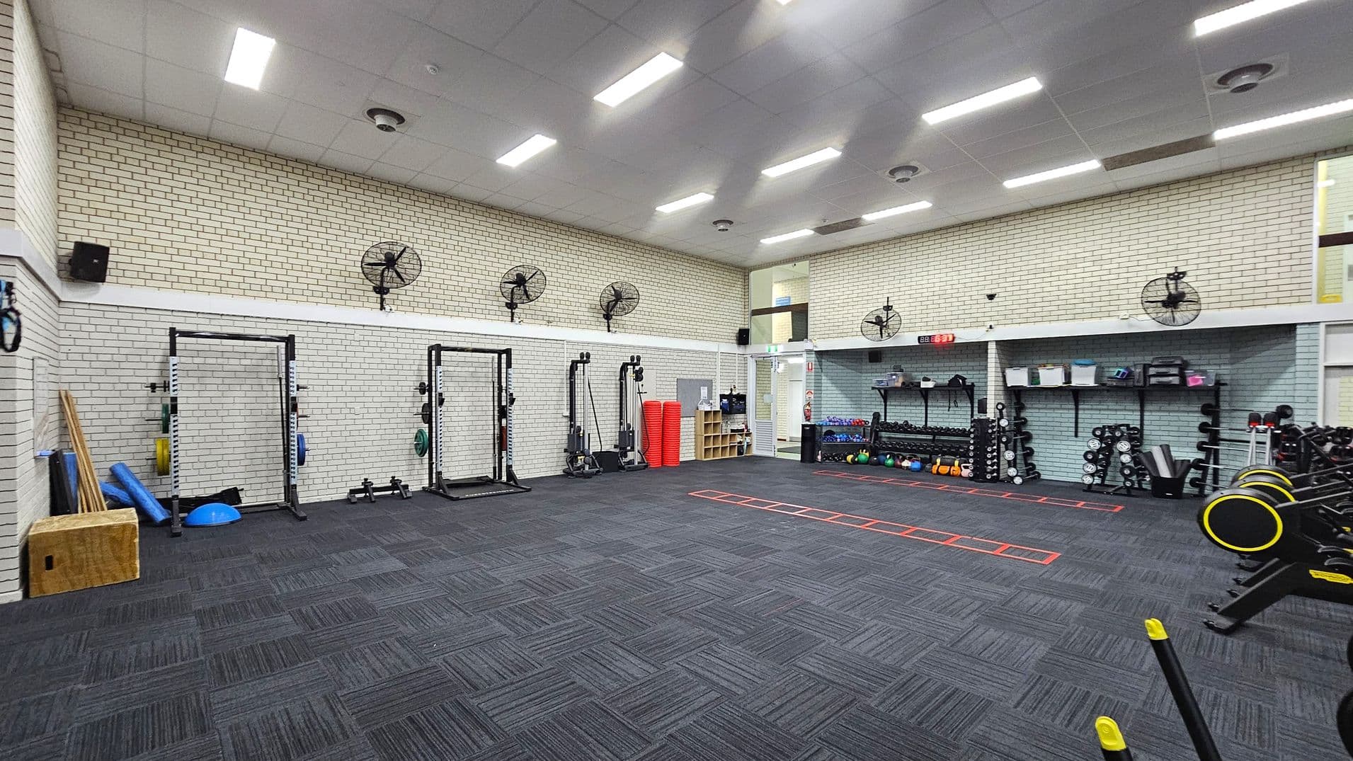 A spacious gym with a dark carpeted floor featuring a mix of grey and purple tones. The left side has exercise equipment like squat racks, weights, and foam rollers. The right side has shelves with kettlebells, dumbbells, and other fitness gear. The walls are light-coloured brick with several wall-mounted fans. The ceiling is high with bright fluorescent lights. A red agility ladder is laid out on the floor.
