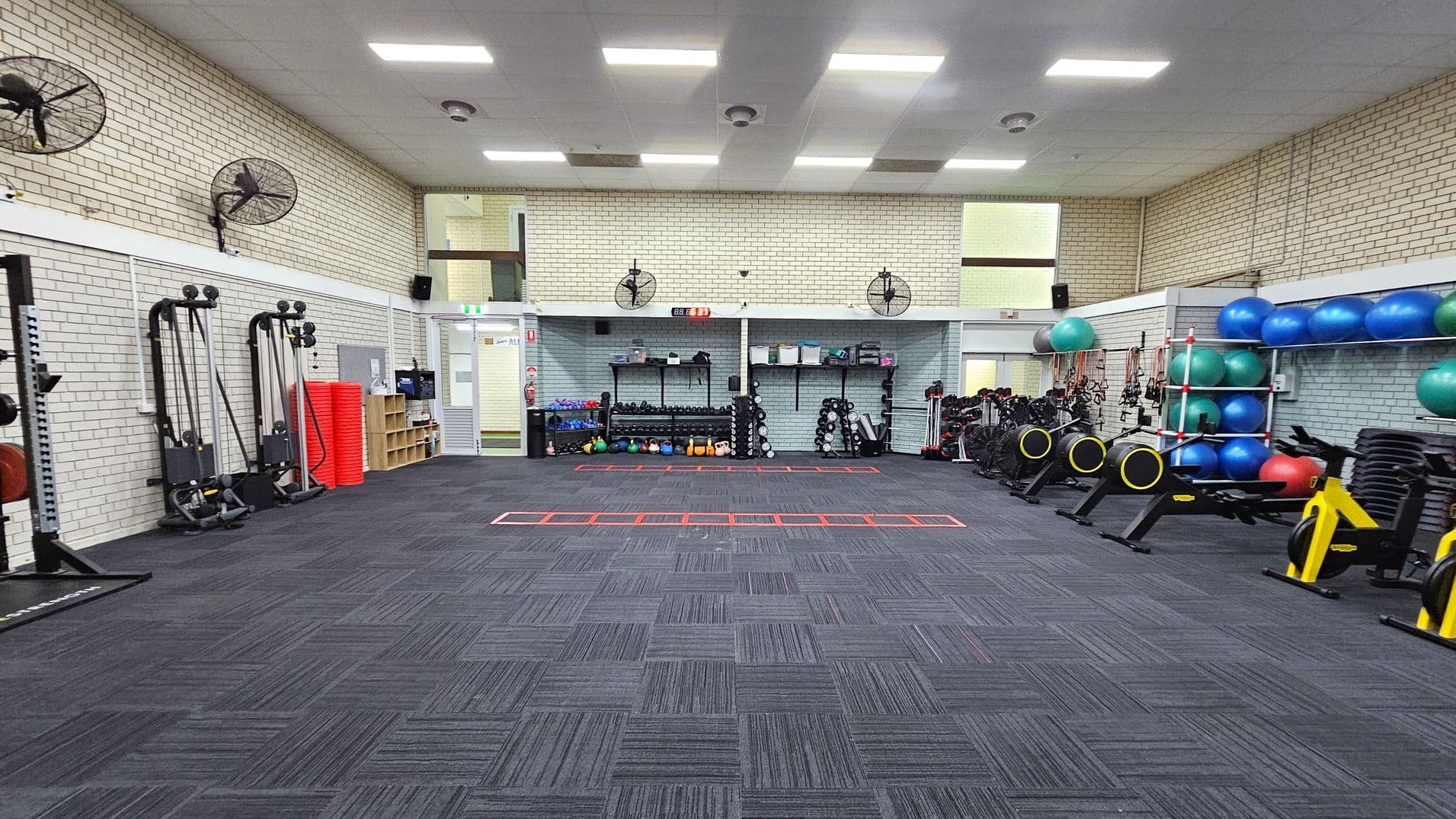 A spacious gym with a dark carpeted floor and a white tiled ceiling. On the left, there are exercise machines and red mats stacked against the wall. The back wall features shelves with kettlebells, dumbbells, and other equipment. On the right, there are exercise bikes and shelves with blue and green exercise balls. Wall-mounted fans are positioned around the room. The walls are light-coloured brick, and there are fluorescent lights on the ceiling.