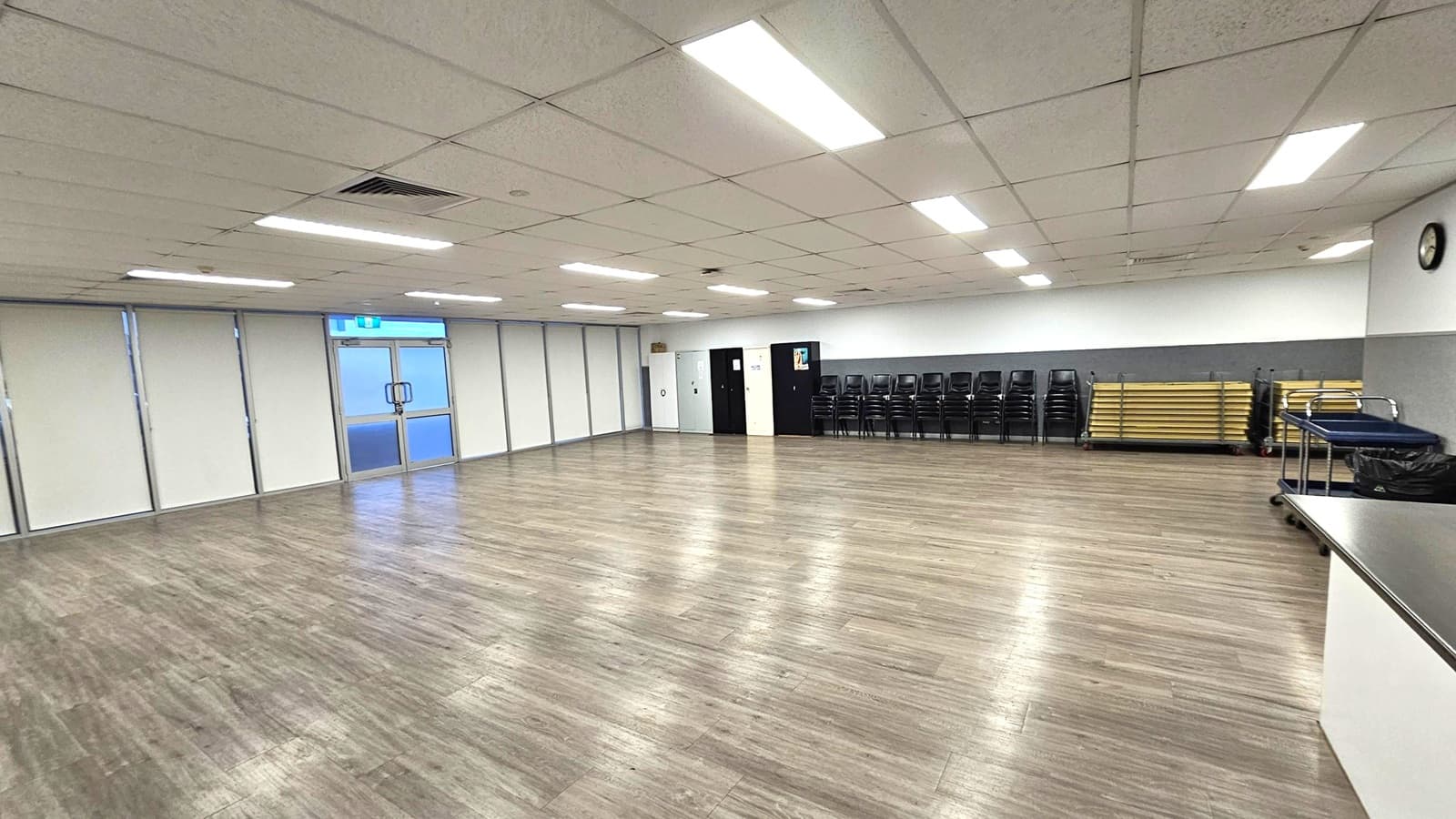 A spacious room with a wooden floor and a grid-patterned ceiling with fluorescent lights. On the left, there are large windows with blinds and a double glass door. The back wall has a stack of black chairs and folded tables. On the right, a clock is mounted on the wall above a counter with a trolley and a bin. The room appears to be a multipurpose space, possibly for meetings or events.