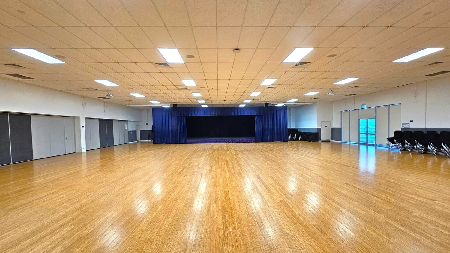A large, empty hall with a polished wooden floor. In the centre, a stage with dark blue curtains is visible. The ceiling is lined with rectangular fluorescent lights. On the left, there are several closed, grey partition doors. On the right, stacked chairs are placed against the wall near a set of glass doors. The walls are plain and light-coloured, contributing to the spacious feel of the room.