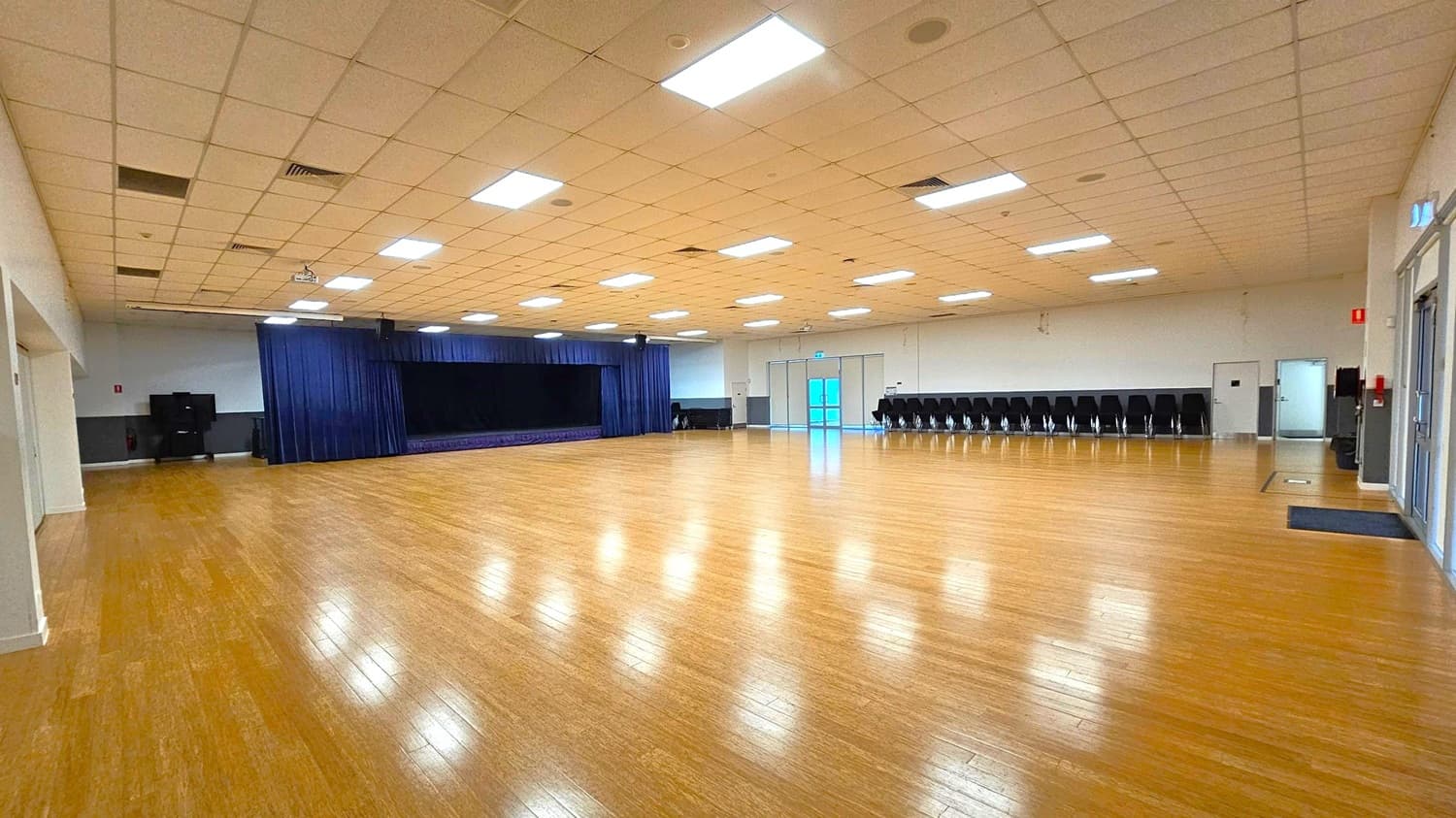 A large, empty hall with a polished wooden floor and a grid-patterned ceiling with fluorescent lights. On the left, a stage with dark blue curtains is visible. The right side features a row of stacked chairs against the wall. At the far end, there are double doors with glass panels. The walls are light-coloured, and there are a few doors and a fire extinguisher on the right.