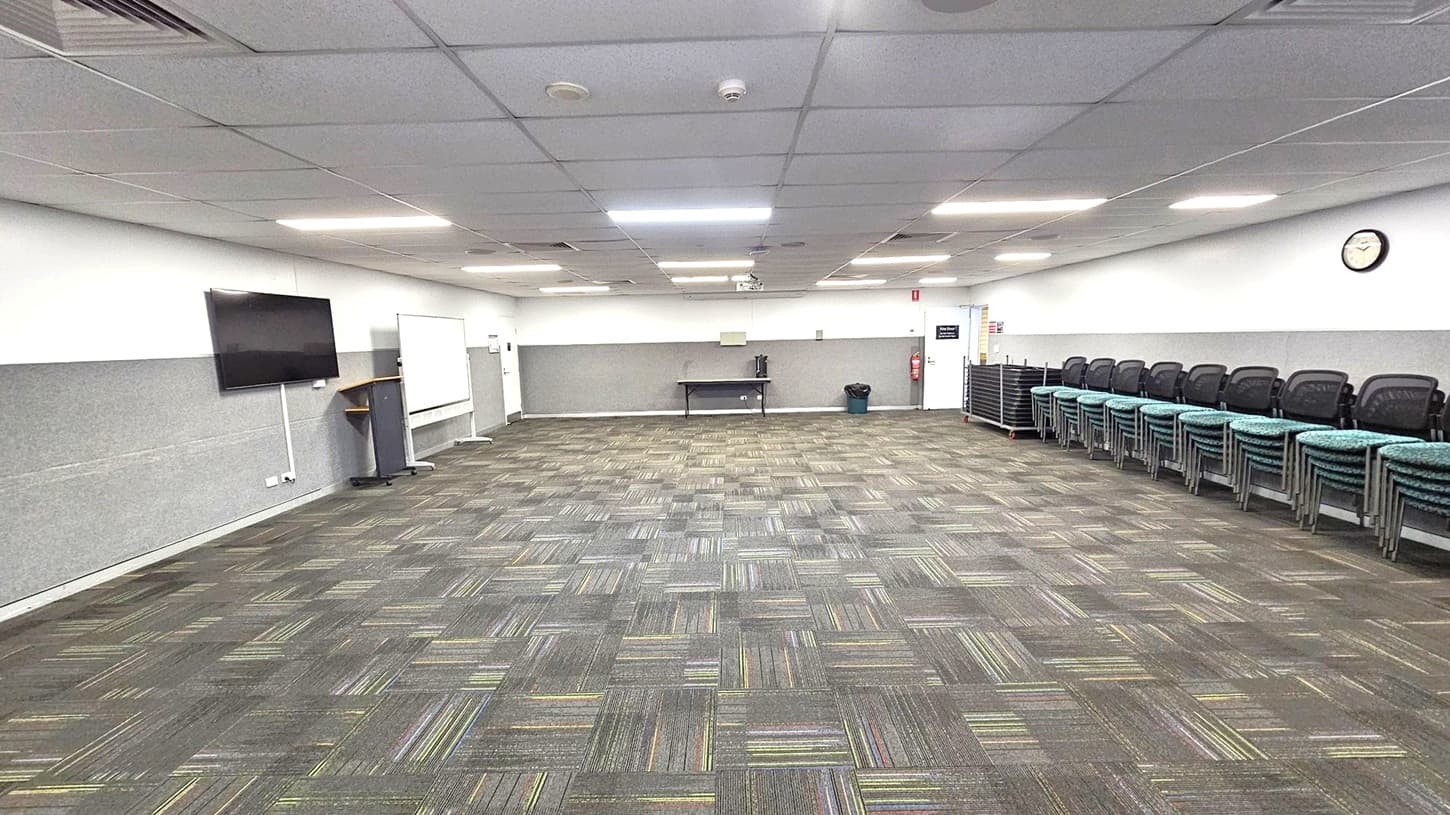 A spacious room with a patterned carpet floor in shades of grey and green. On the left, a wall-mounted TV and a lectern are visible. In the centre, a whiteboard and a table with a kettle are against the wall. On the right, a stack of chairs is neatly arranged, and a clock is mounted on the wall. The ceiling is fitted with fluorescent lights and air vents.