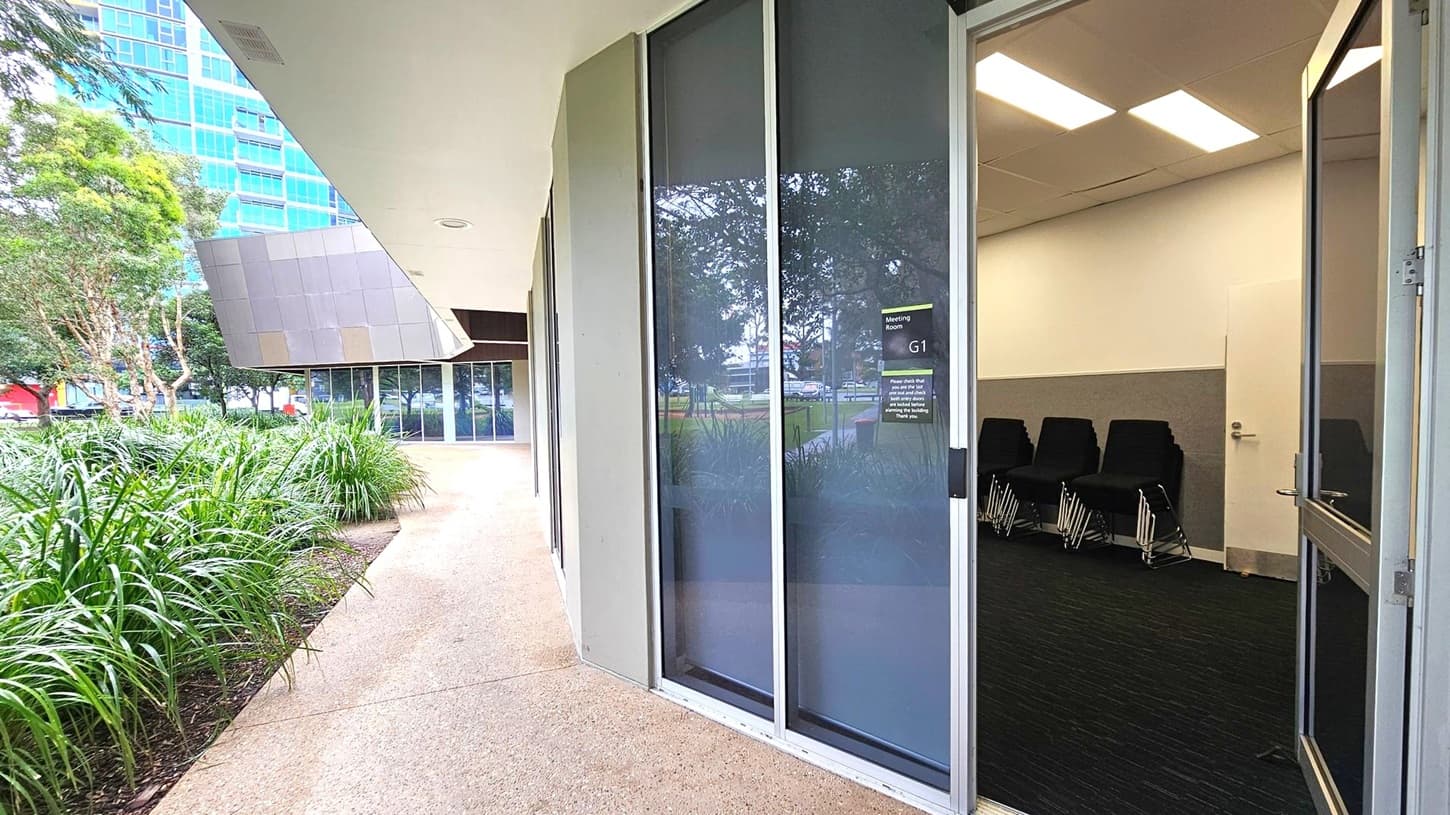 A modern building exterior with a pathway lined by lush green plants on the left. The building has large glass windows and an open door leading to a meeting room labelled "G1" on the right. Inside, several black chairs are stacked against a wall. The ceiling has rectangular fluorescent lights. In the background, a tall building with blue-tinted windows is visible. The floor outside is a textured concrete surface.