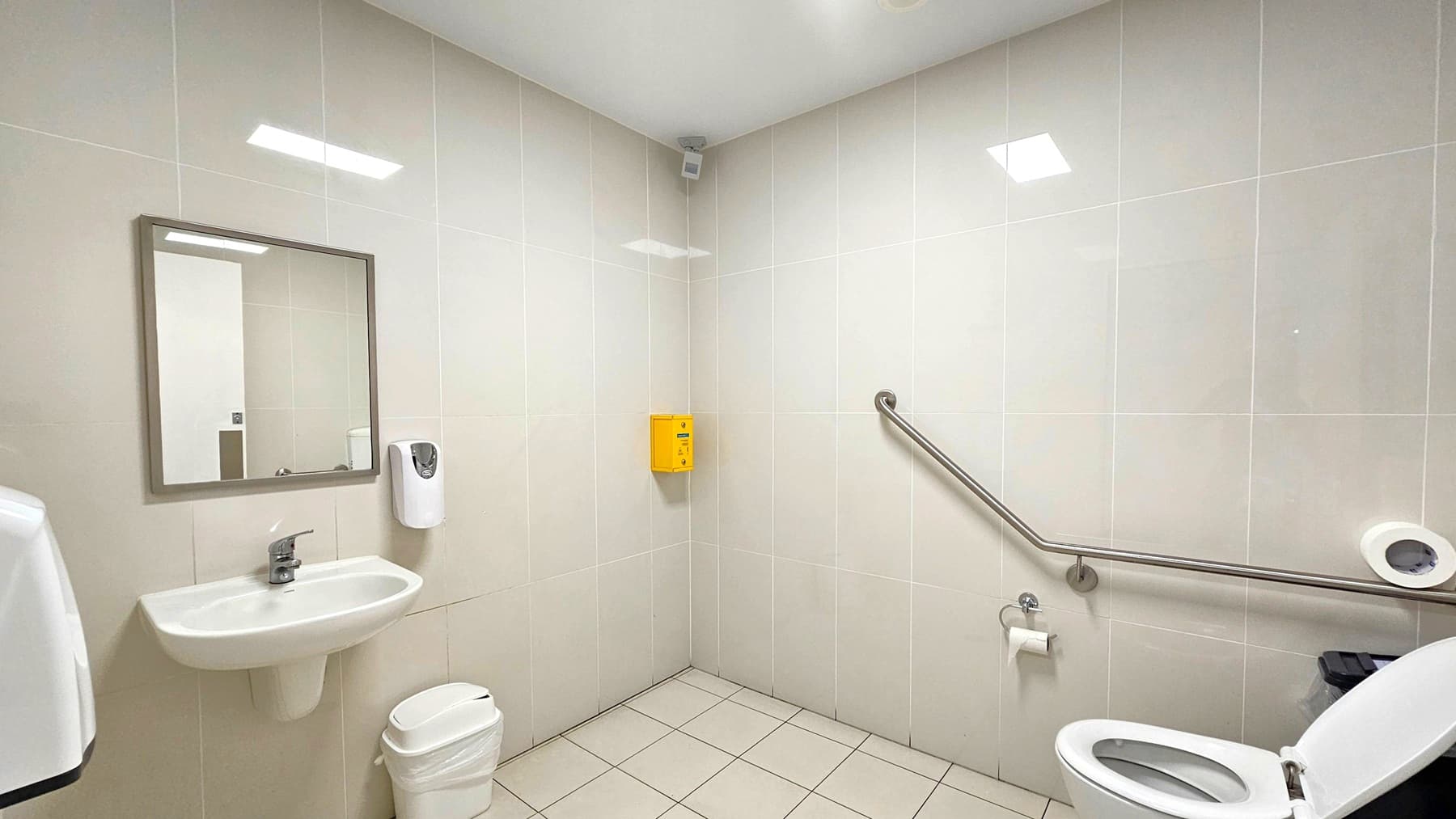 A clean, accessible bathroom with white tiled walls and a light-coloured tiled floor. On the left, there's a wall-mounted sink with a mirror above it and a soap dispenser to the right. Below the sink is a small waste bin. On the right, a toilet with an open lid is equipped with a grab rail for accessibility. A toilet paper holder is mounted on the wall. A yellow box is fixed in the corner above the grab rail. The ceiling is plain white with recessed lighting.