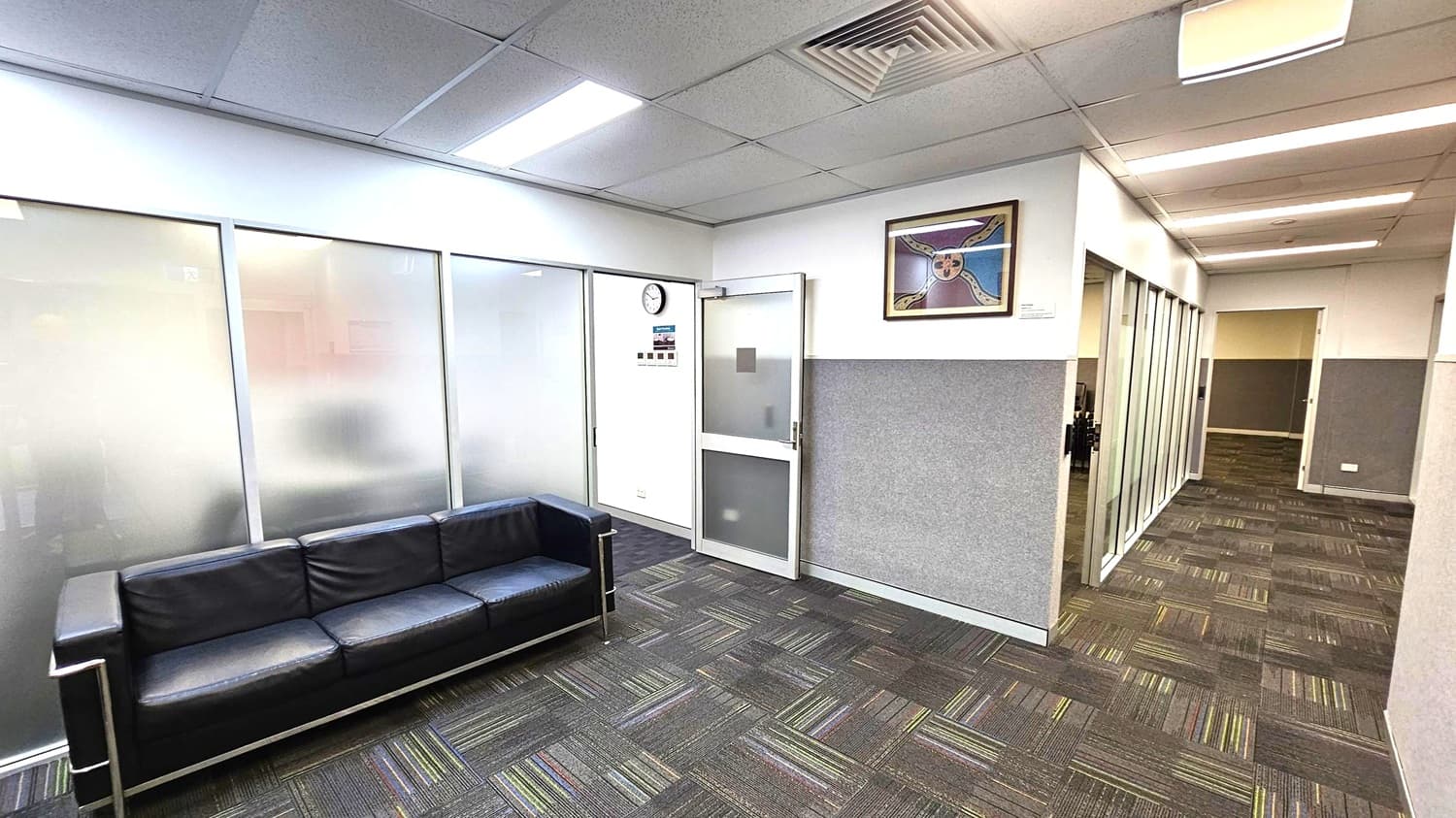 A modern office interior with a black leather sofa on the left against frosted glass panels. The floor is covered with patterned carpet tiles in shades of grey and green. On the right, a corridor with glass partitions leads to other rooms. A wall clock and a colourful framed artwork are mounted on the wall above a door. The ceiling features recessed lighting and air vents.