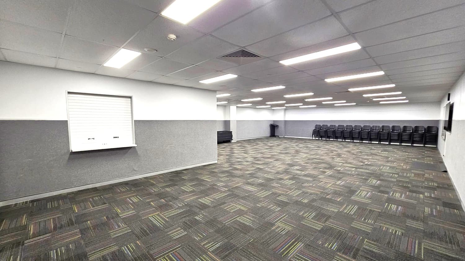 A large, empty room with a patterned carpet featuring grey and green tones. The ceiling is lined with rectangular fluorescent lights. On the left wall, there's a closed shutter window. The right side has a stack of black chairs lined up against the wall. In the background, more chairs are stacked, and a bin is visible. The walls are two-toned, with a darker shade on the lower half.