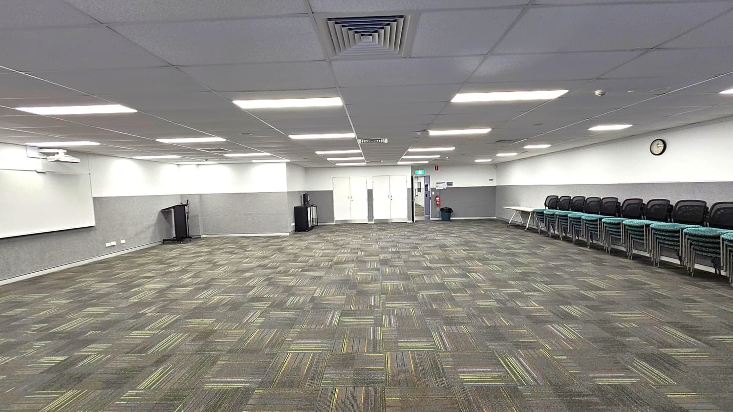 A spacious room with a patterned carpet in shades of green and grey. On the left, there's a whiteboard and a podium. The back wall features double doors and a small cabinet. On the right, a row of stacked chairs lines the wall beneath a clock. The ceiling is fitted with fluorescent lights and ventilation panels.