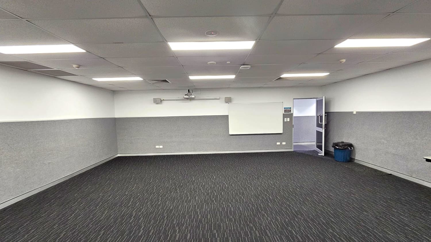 A room with a dark carpeted floor featuring a subtle linear pattern. The walls are light grey with a whiteboard mounted on the centre of the far wall. Above the whiteboard, a projector is installed. On the right, an open door leads to another area, and a blue bin is placed nearby. The ceiling is fitted with rectangular fluorescent lights, evenly spaced across the room.