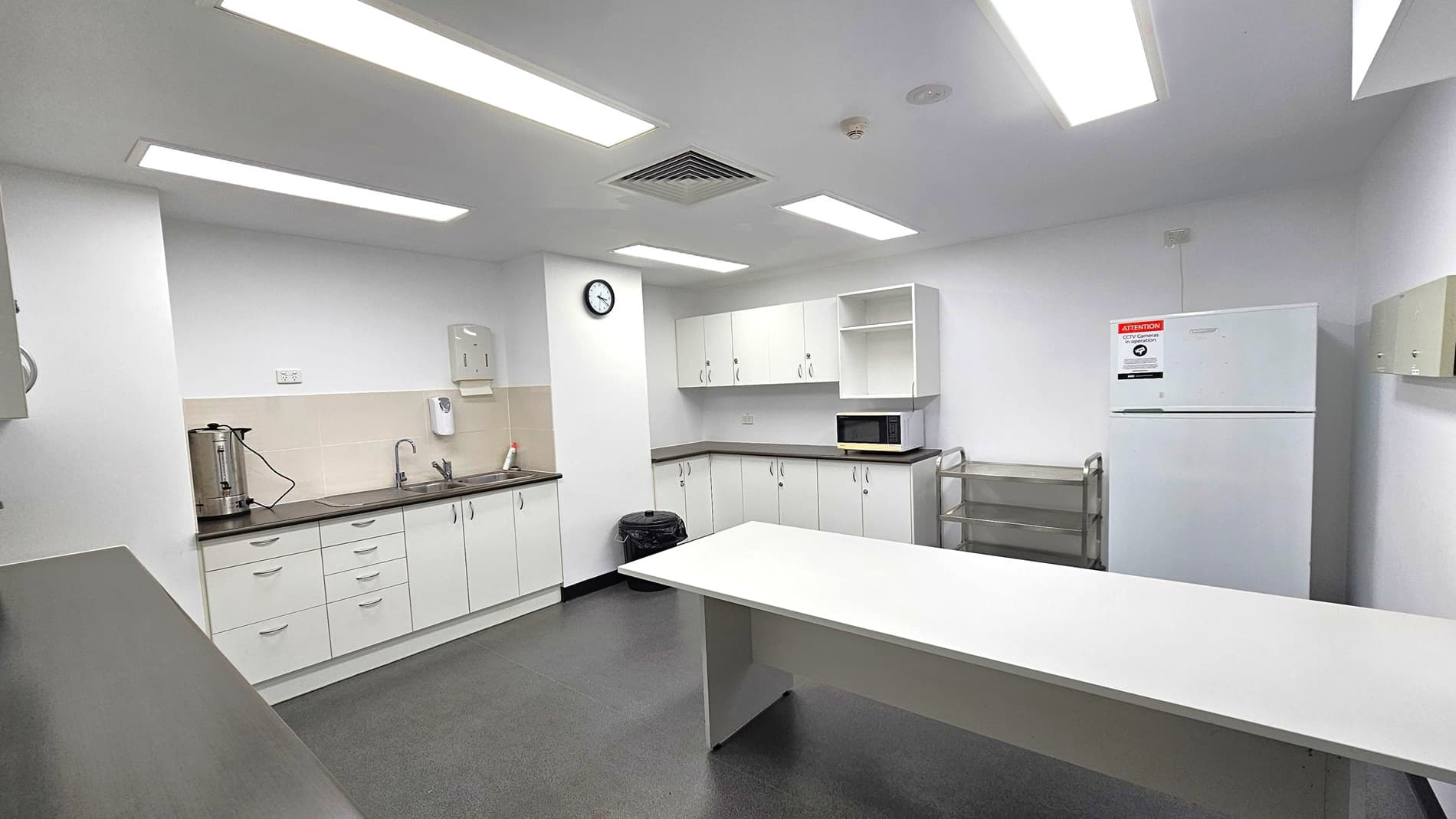 A clean, modern kitchen with white cabinetry and a dark countertop. On the left, a sink with a kettle and soap dispenser is visible. A wall clock is mounted above. In the centre, a white table stands on a dark floor. On the right, a microwave sits on a counter, with a refrigerator nearby. The ceiling has several rectangular fluorescent lights and a vent. A small metal trolley is positioned next to the fridge.