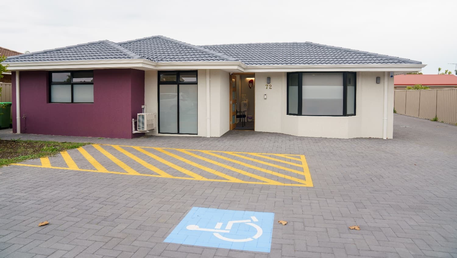 A closer look at the cream building with a grey, tiled roof with a feature wall to the left in purple. The front door is timber with four glass panels and is open with a glimpse of a chair is visible inside. A cream colourbond fence sits t the right and a red roof is visible to the right and behind this business. An accessible car parking space is directly adjacent to the front entrance.