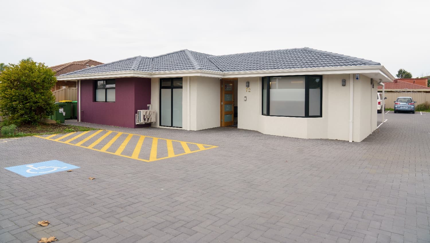 Another angle of a view of the cream building with a grey, tiled roof with a feature wall to the left in purple. The front door is timber with four glass panels. To the left is a white sign with 'Periodontics & Implants'. To the right is a glimpse of a small, white van and a small grey vehicle parked behind the building. An accessible car parking space is directly adjacent to the front entrance. To the left of this buiding are the rubbish bins and a glimpse of the building located next door.