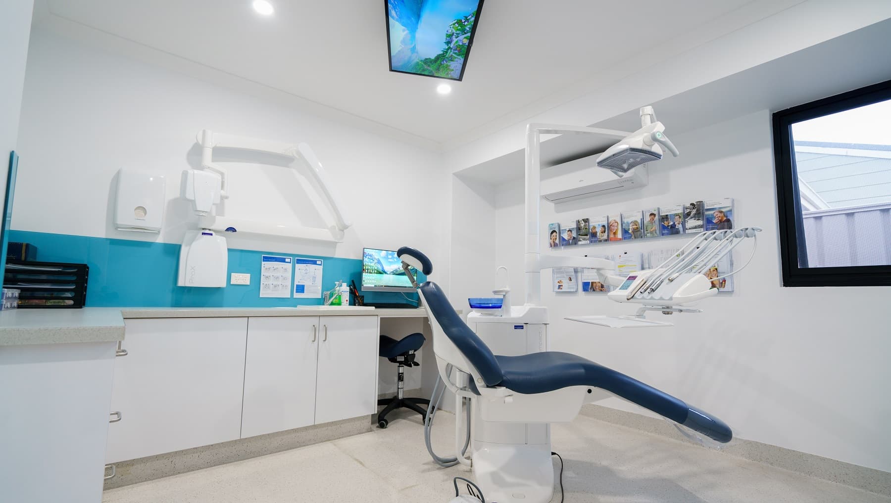 The walls of the examination room are white with sky-blue accents. The clinic has a reclining examination chair in the middle of the room where the basic equipment used by periodontists are attached, such as the bright light used during the examination, spittoon, and suction. Sunlight is visible through the glass window. There are magazine racks mounted on the wall on the right side of the room where reading materials are displayed.
