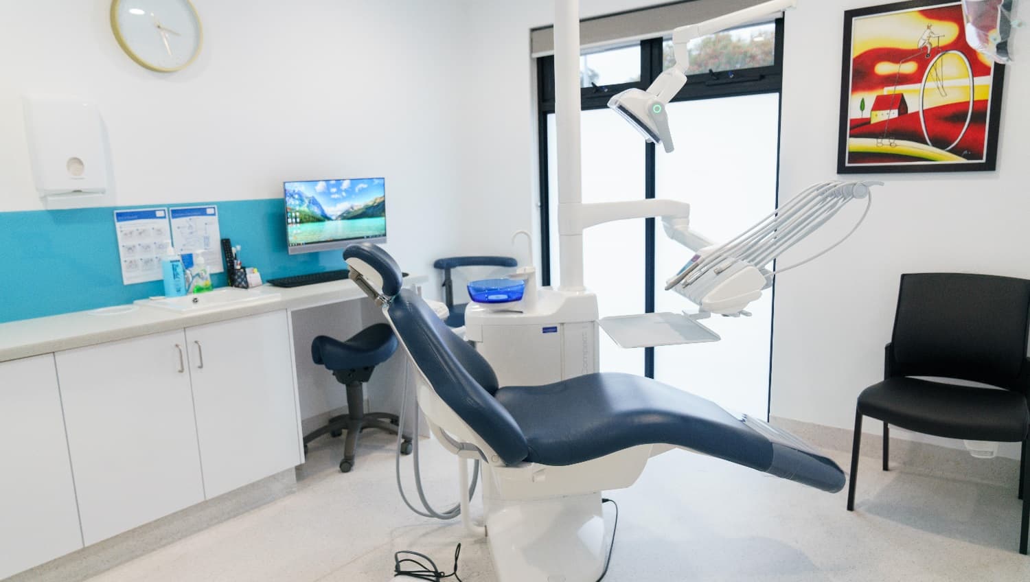 This view of an examination room shows a painting posted on the wall. The artist used bright and vibrant shades of yellow and red. It has a dark blue reclining examination chair that looks very comfortable. This chair is attached to the equipment used by dentists/periodontists. A wall clock is mounted on the wall and below it is a rectangular white tissue dispenser. A black chair is also kept near the examination chair and dental equipment.
