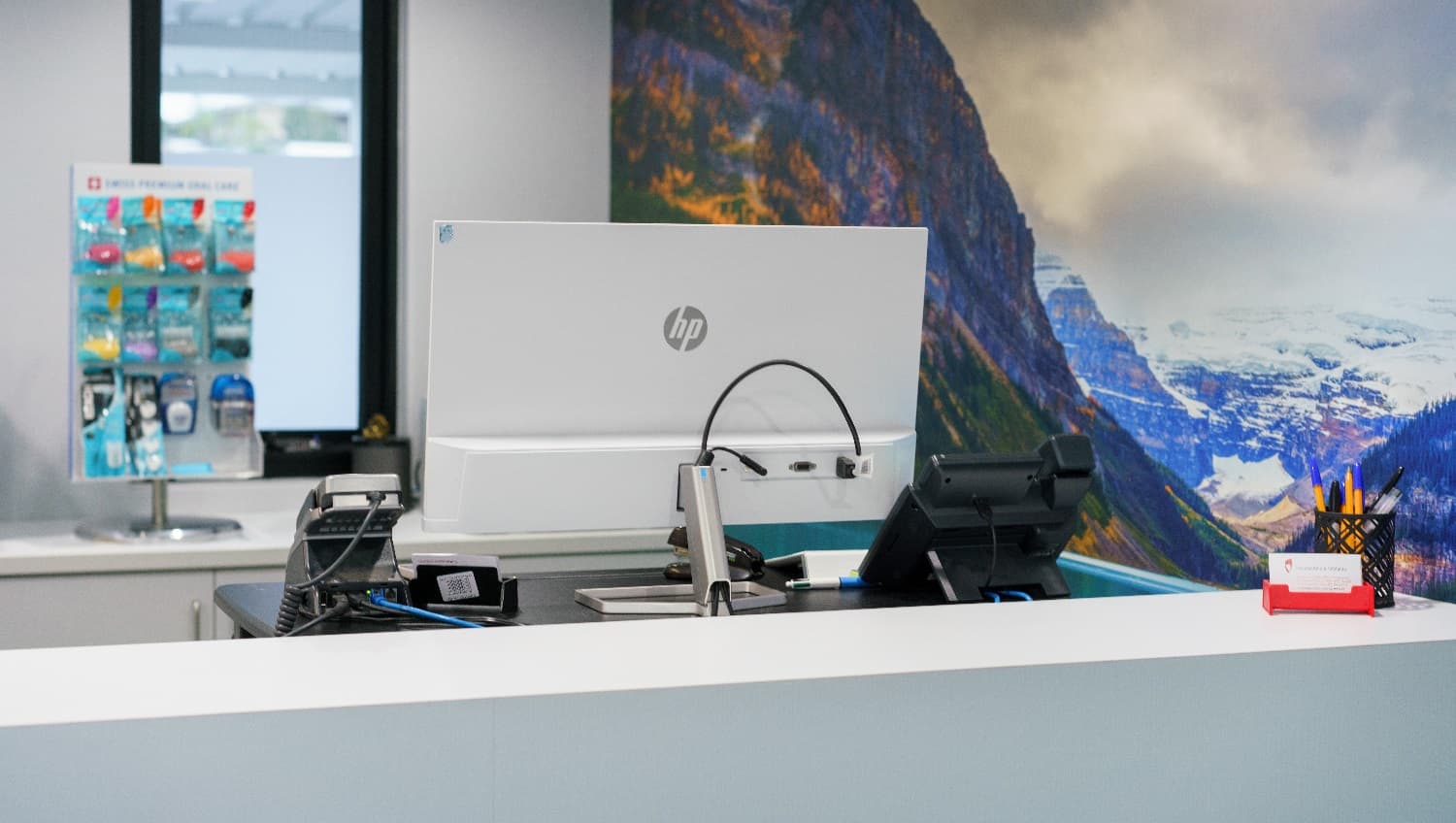 A closer look of the Periodontics & Implants clinic’s desk that has the desktop computer and telephone unit for use by the staff. The clinic’s business cards and pens are also available on the corner of the desk that’s close to the wall-sized landscape. There are dental health items that are for sale on the back counter.