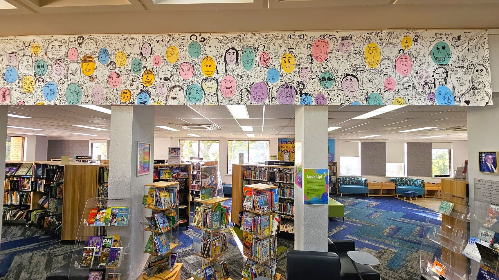 A library interior with a colourful mural of hand-drawn faces on the upper wall. Bookshelves filled with books are in the centre and left, with a display of children's books in the foreground. The floor is carpeted in a blue and green pattern. Large windows in the background allow natural light in, and there are blue cushioned seating areas. A framed picture is on the right wall, and a poster is on a pillar. The ceiling has fluorescent lights.