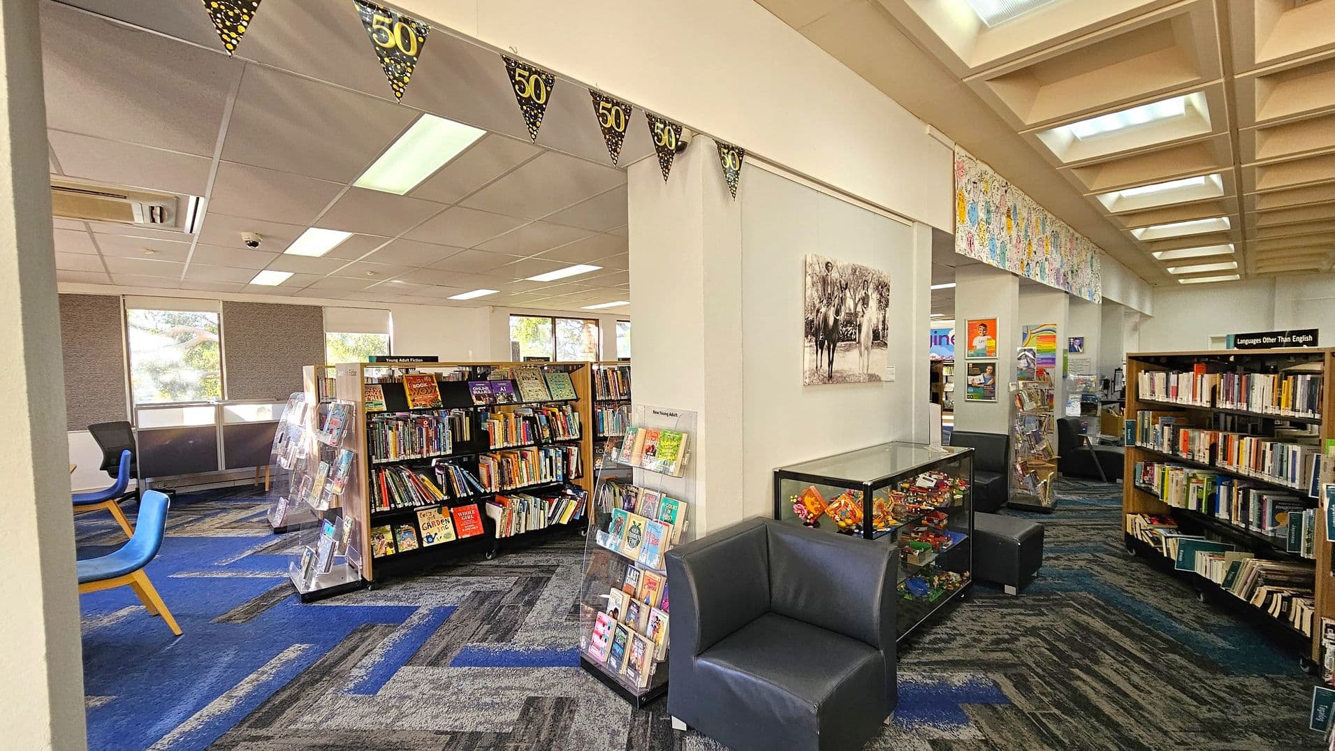A library interior with a blue and grey patterned carpet. On the left, there are bookshelves filled with books and a few blue chairs near windows. In the centre, a display rack holds various books and magazines. A black armchair is positioned next to a glass display case containing colourful items. On the right, more bookshelves are labelled "Languages Other Than English." The ceiling features recessed lighting and decorative banners with "50" hang across the room.