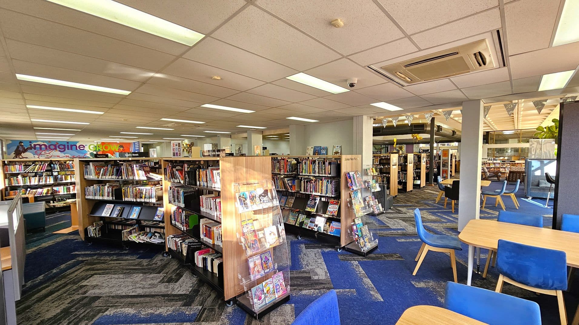 A library interior with bookshelves filled with books in the centre and left. A colourful sign with the words "imagine" and "GROW" is visible on the left wall. On the right, there are tables with blue chairs. The floor is carpeted in a blue and grey pattern, and the ceiling has recessed lighting and an air conditioning unit. In the background, more bookshelves and seating areas are visible, along with some decorative bunting.