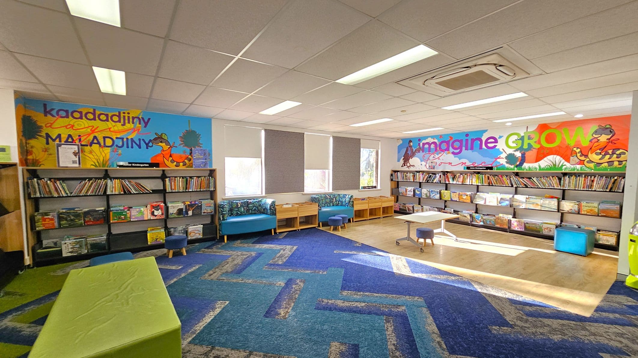 A colourful library space with bookshelves lining the walls. On the left, a mural with the words "kaadadjiny" and "maladjiny" above a bookshelf filled with books. In the centre, there are blue cushioned benches and small tables with stools. The right wall features another mural with the words "imagine" and "GROW" above more bookshelves. The floor has a blue patterned carpet, and the ceiling is fitted with fluorescent lights and an air conditioning unit.