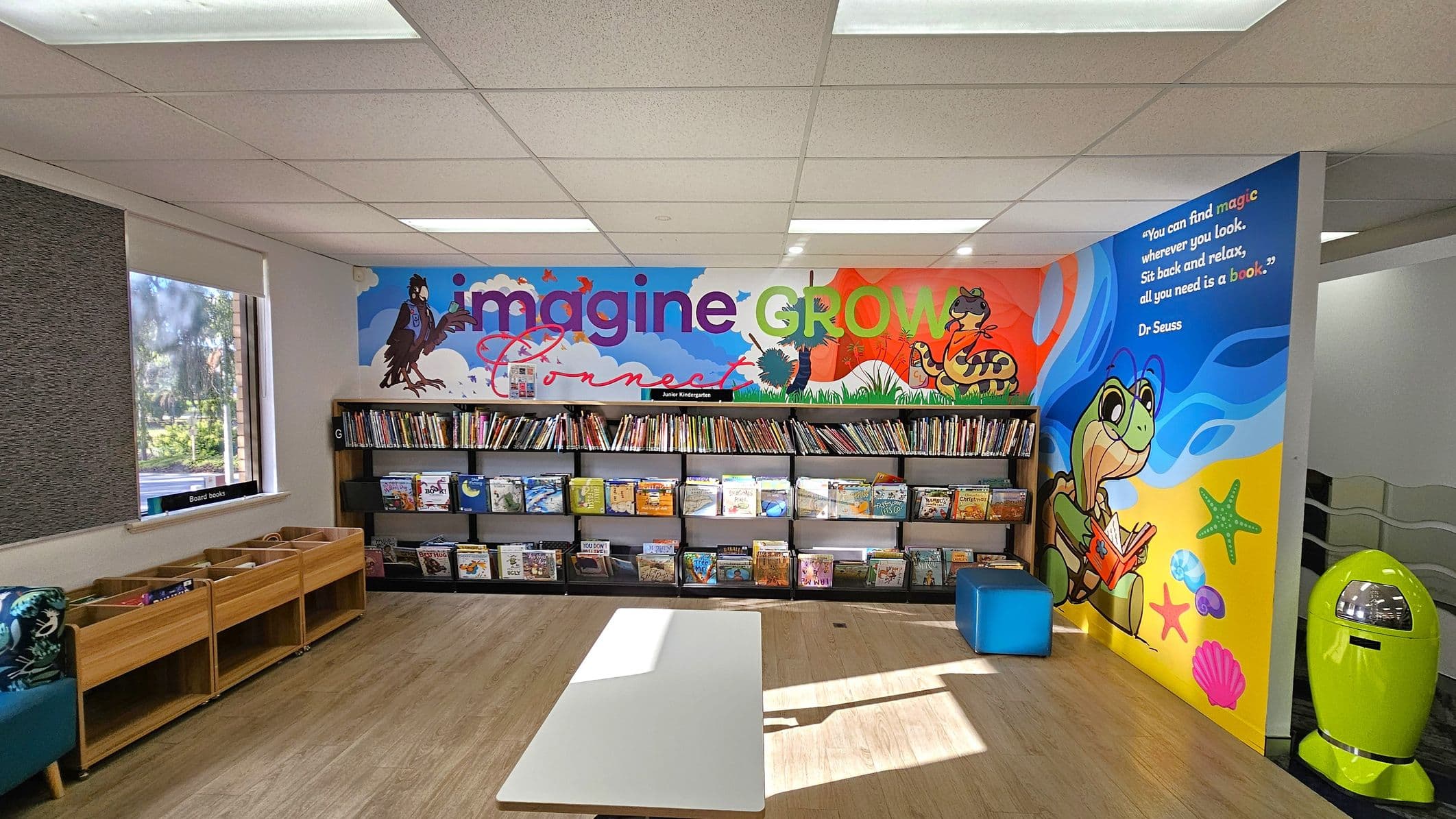 A colourful children's library area with a vibrant mural on the back wall featuring the words "imagine," "GROW," and "Connect," along with cartoon animals. Below the mural, there are shelves filled with children's books. On the left, wooden book bins are placed under a window. On the right, a bright green bin and a blue cushioned seat are visible. The floor is wooden, and the ceiling has white panels with fluorescent lights.