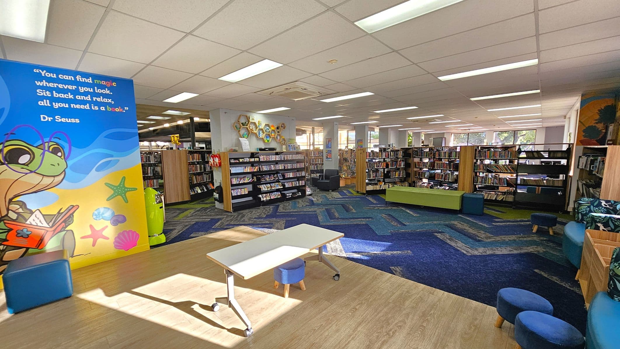 A library interior with a colourful mural on the left featuring a cartoon turtle and a quote by Dr. Seuss. In the centre, there are bookshelves filled with books, and a small white table with a blue stool on a wooden floor. The floor transitions to a blue patterned carpet. On the right, there are more bookshelves and seating, including a green bench and blue cushioned stools. The ceiling is lined with fluorescent lights, and large windows in the background allow natural light in.